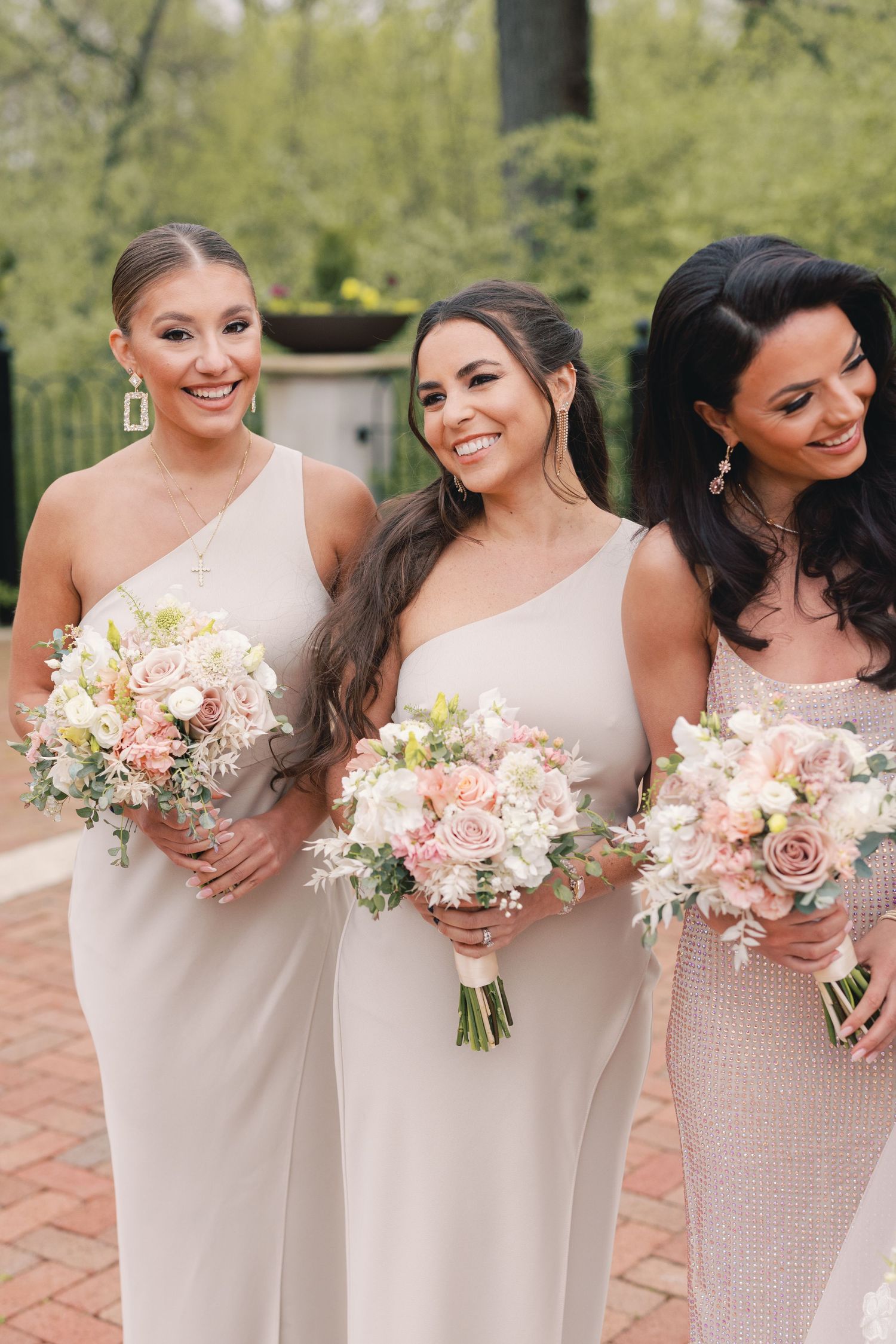 Three bridesmaids in champagne one-shoulder gowns holding blush and white floral bouquets smiling outdoors.