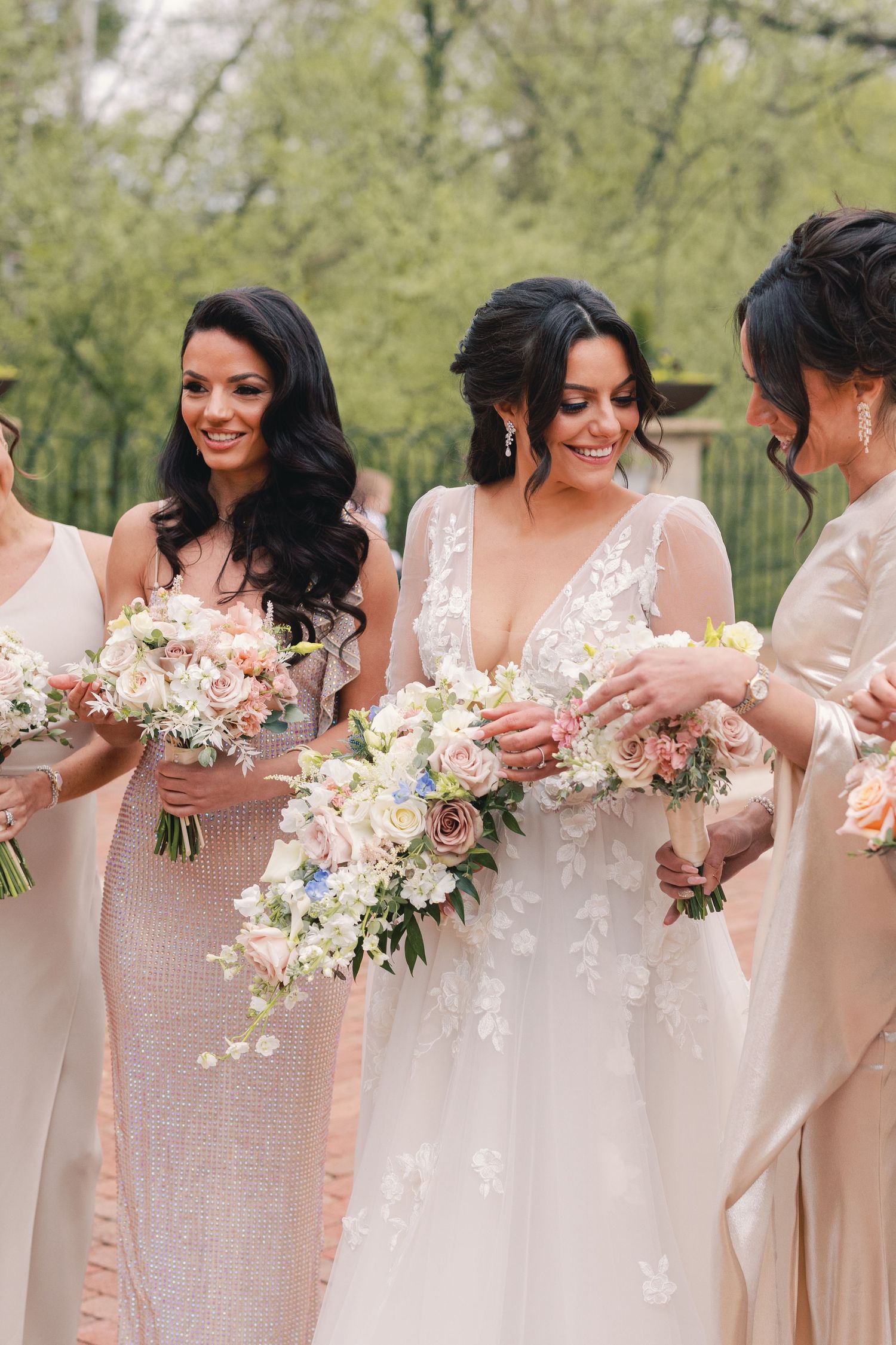 Bride in lace gown laughing with bridesmaids in champagne dresses holding floral bouquets.