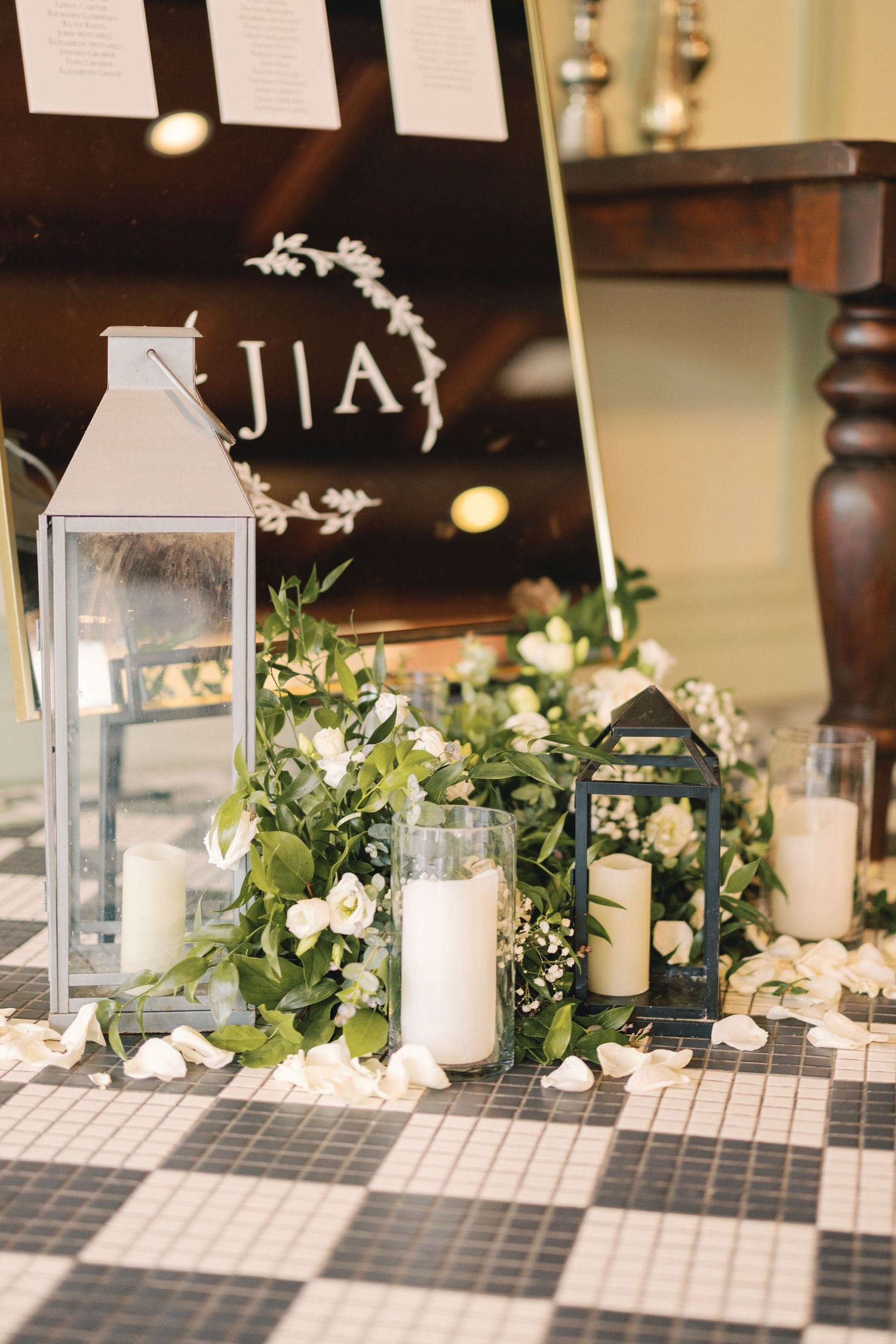 Elegant wedding entrance with white lanterns, candles, greenery, and monogram JIA sign on black tile floor.