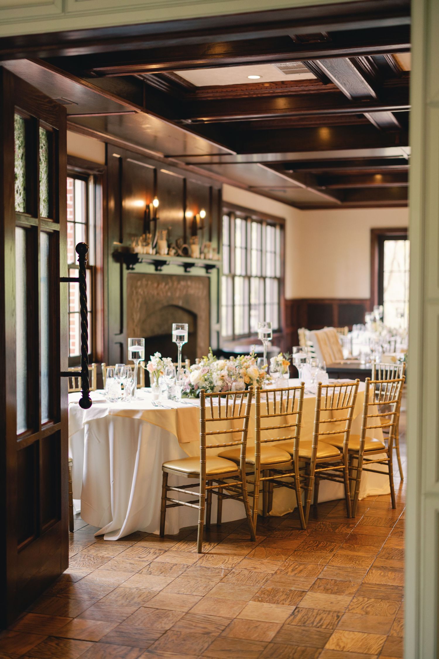 Elegant wedding reception room with dark wood beams, gold chiavari chairs, white linens, and fireplace.