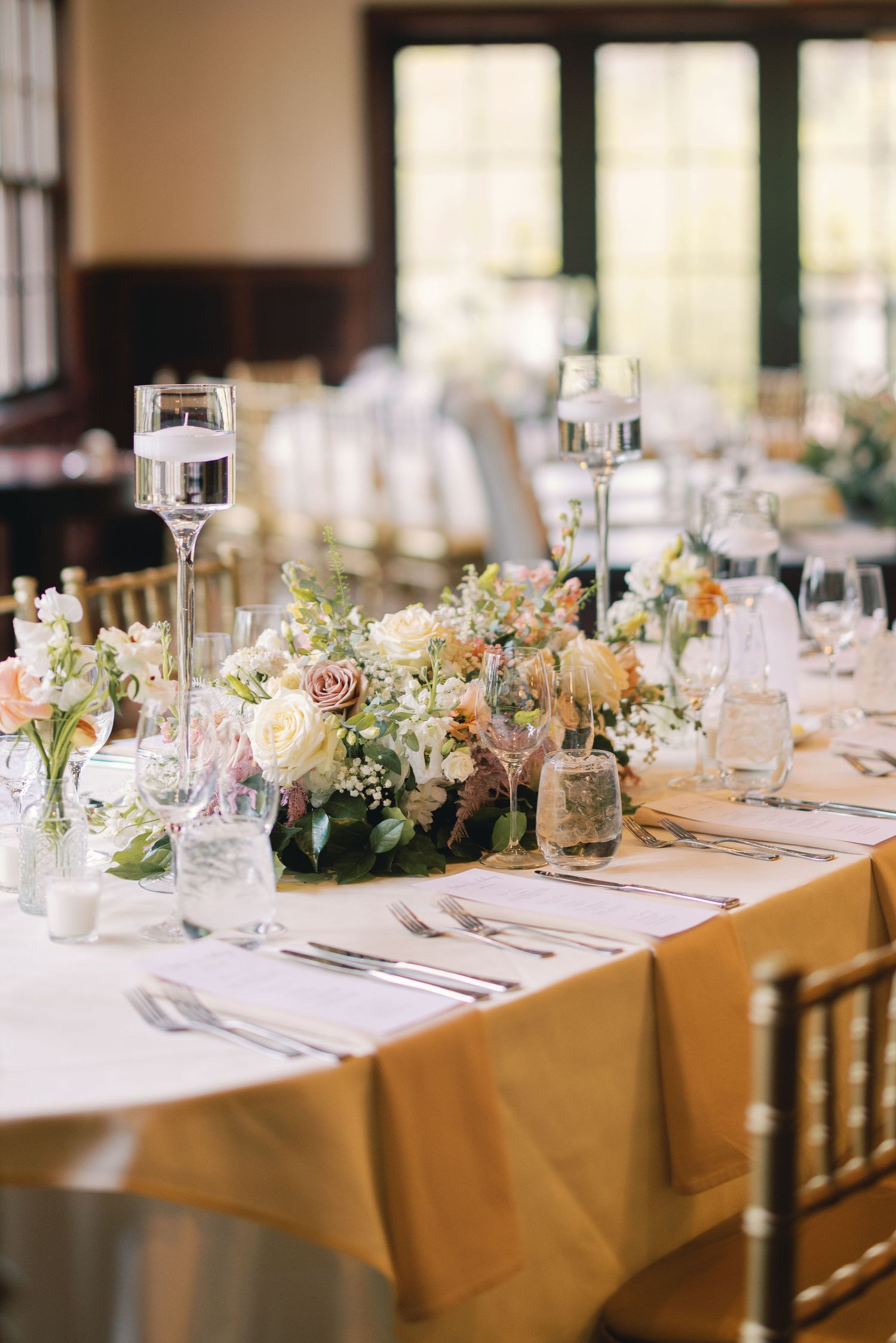 Wedding reception table with gold linens, crystal candleholders, and pastel floral centerpieces in a bright venue.