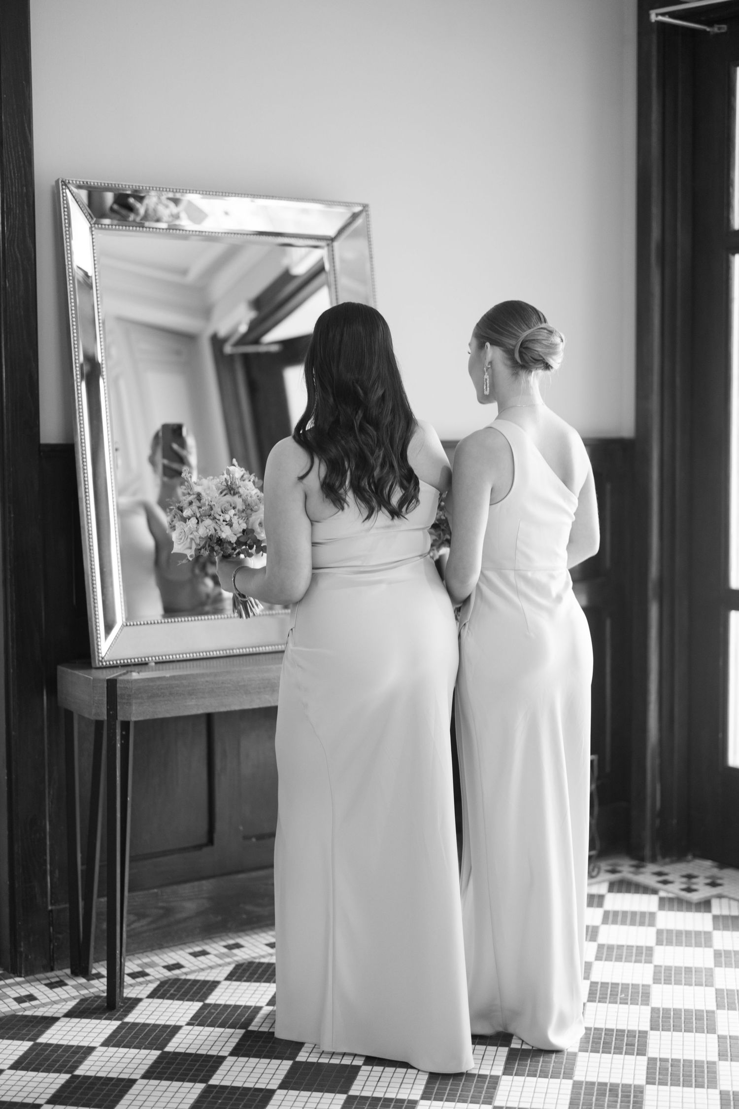 Black and white photo of two bridesmaids in white gowns standing before a large mirror.