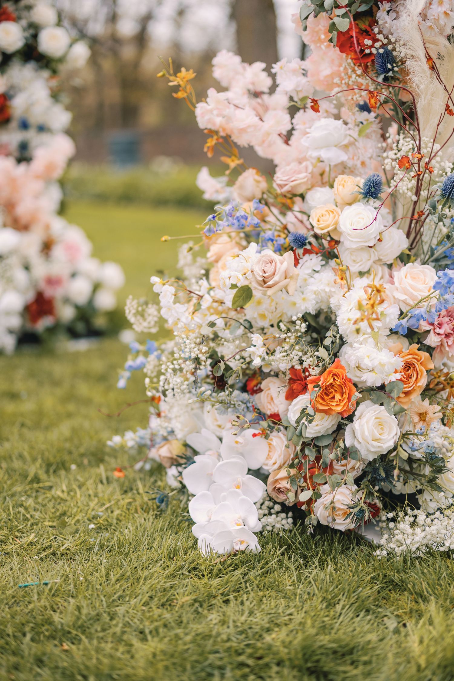 Lush wedding ceremony arch base with white, peach, orange, and blue flowers on green grass.