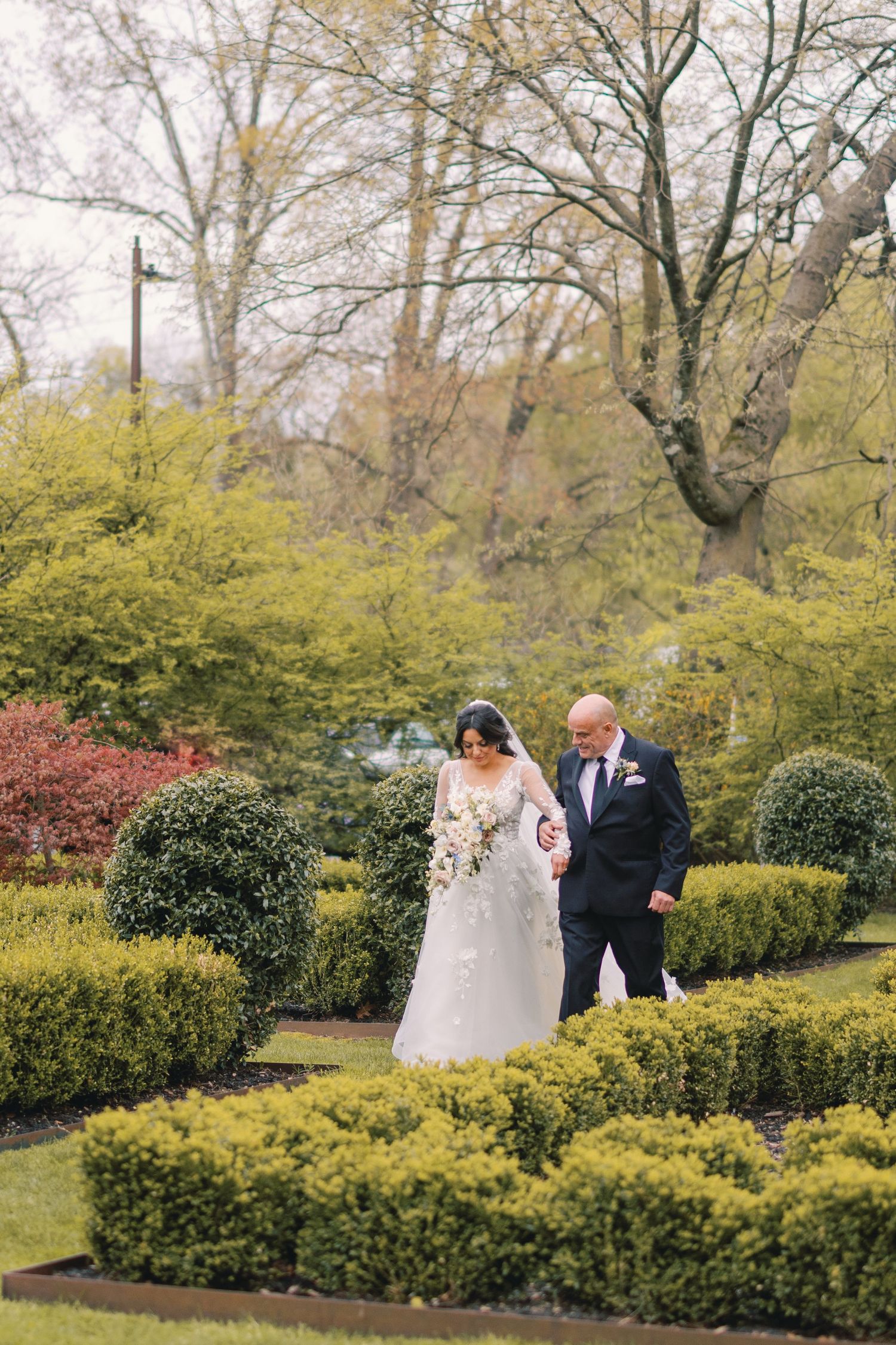 Bride in lace gown holding bouquet walks with escort through manicured garden hedges at outdoor wedding.