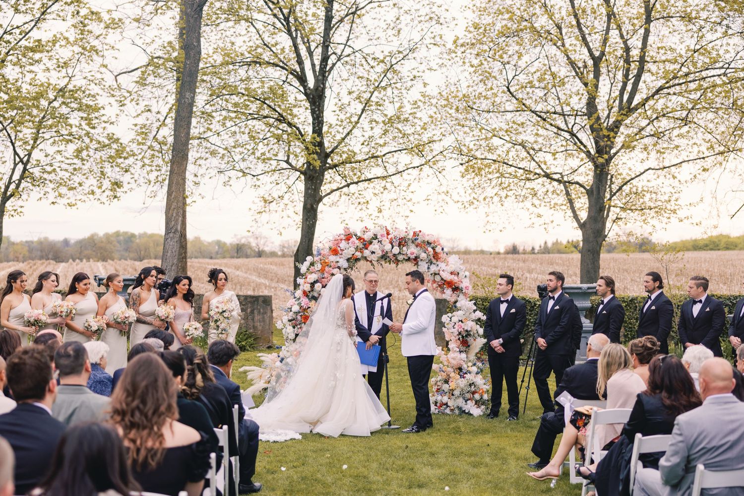 Couple wed under floral arch at outdoor garden ceremony surrounded by guests on sunny day.