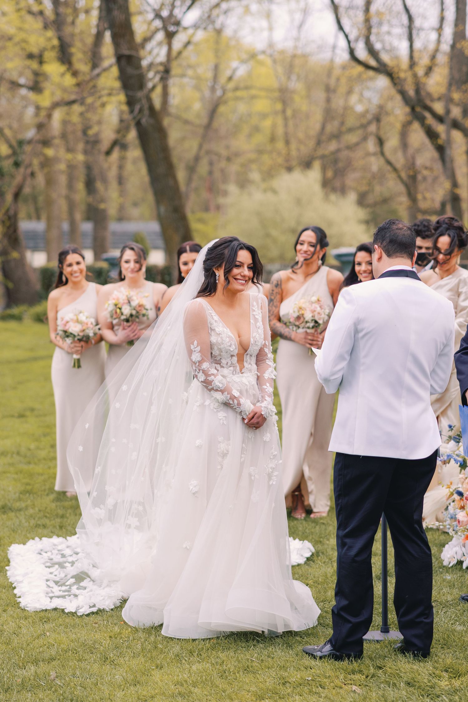 Bride in lace gown with veil stands with bridesmaids in champagne dresses at outdoor ceremony.