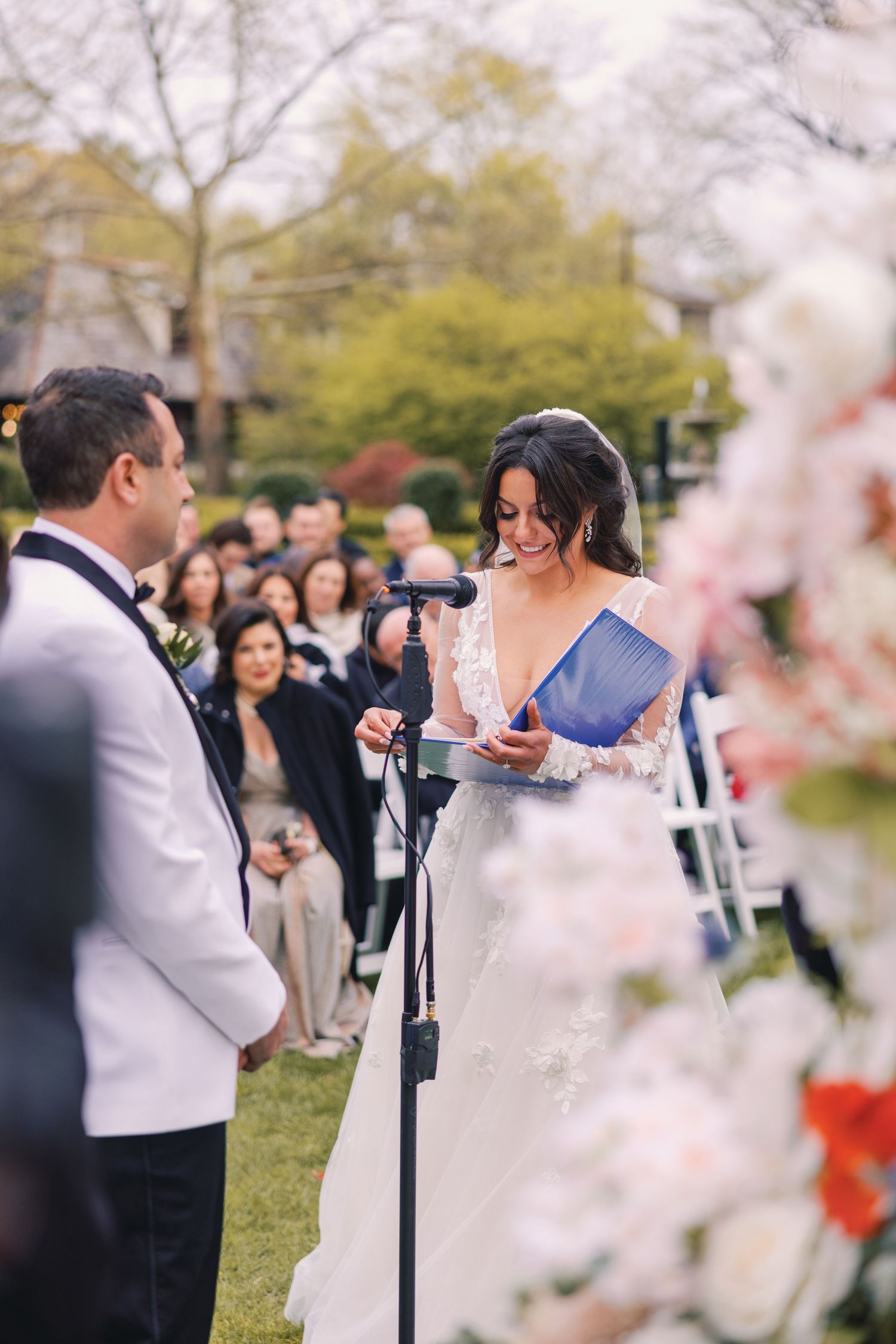 Smiling bride reads vows from blue booklet at outdoor wedding ceremony microphone stand.