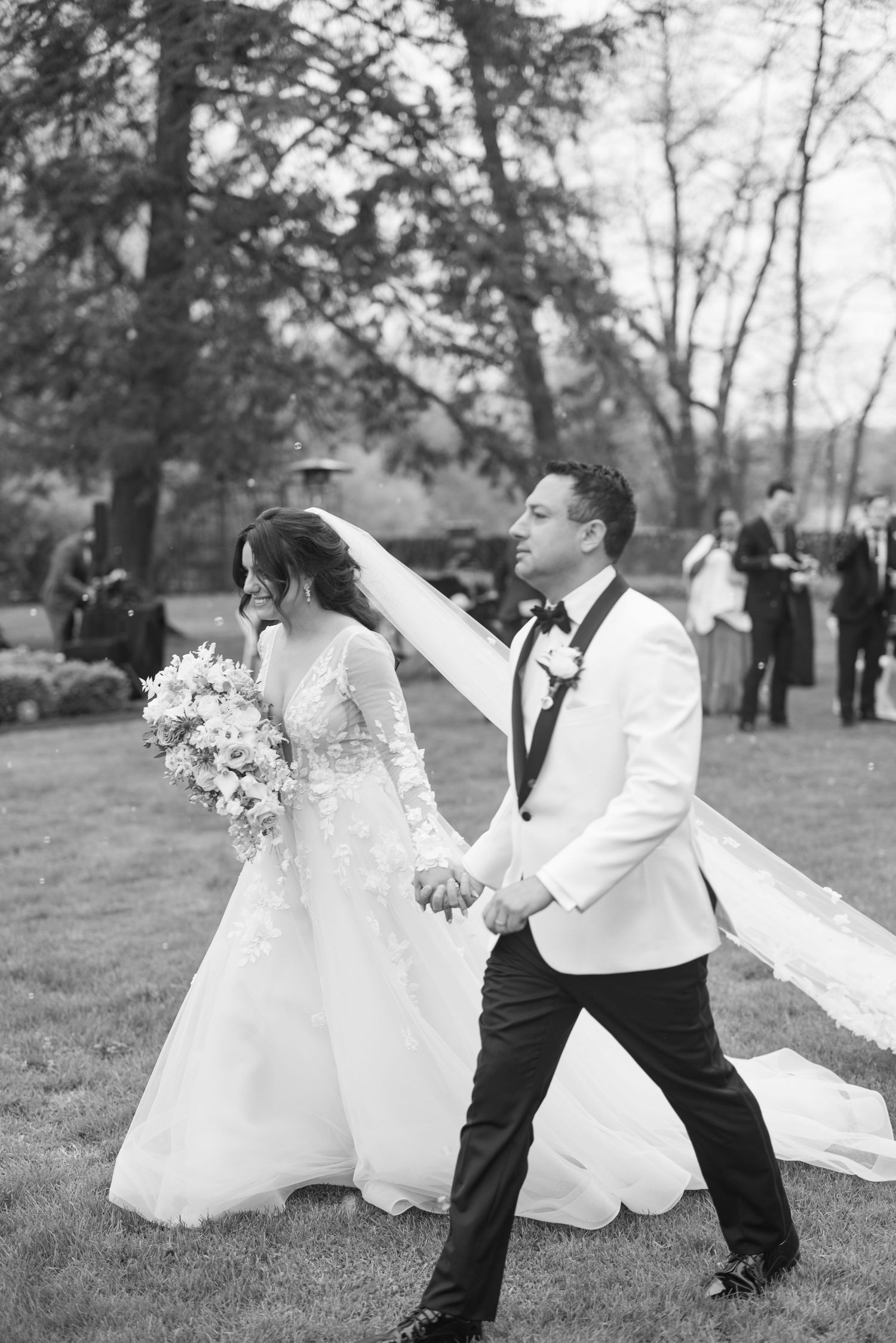 Black and white photo of newlyweds walking hand in hand across lawn after wedding ceremony.