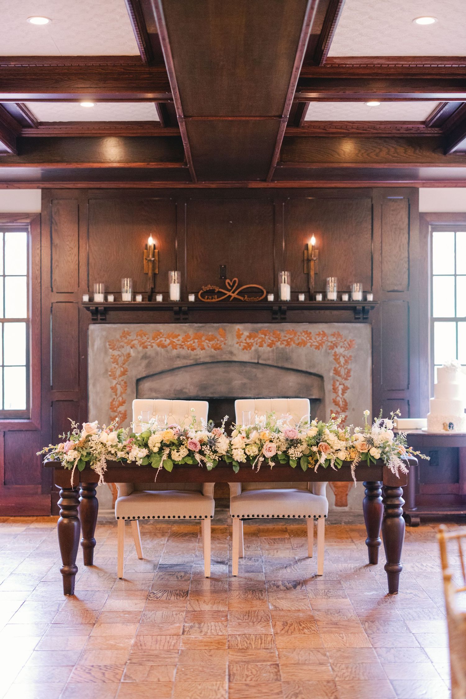 Sweetheart table with floral arrangements set before stone fireplace in rustic wedding venue.