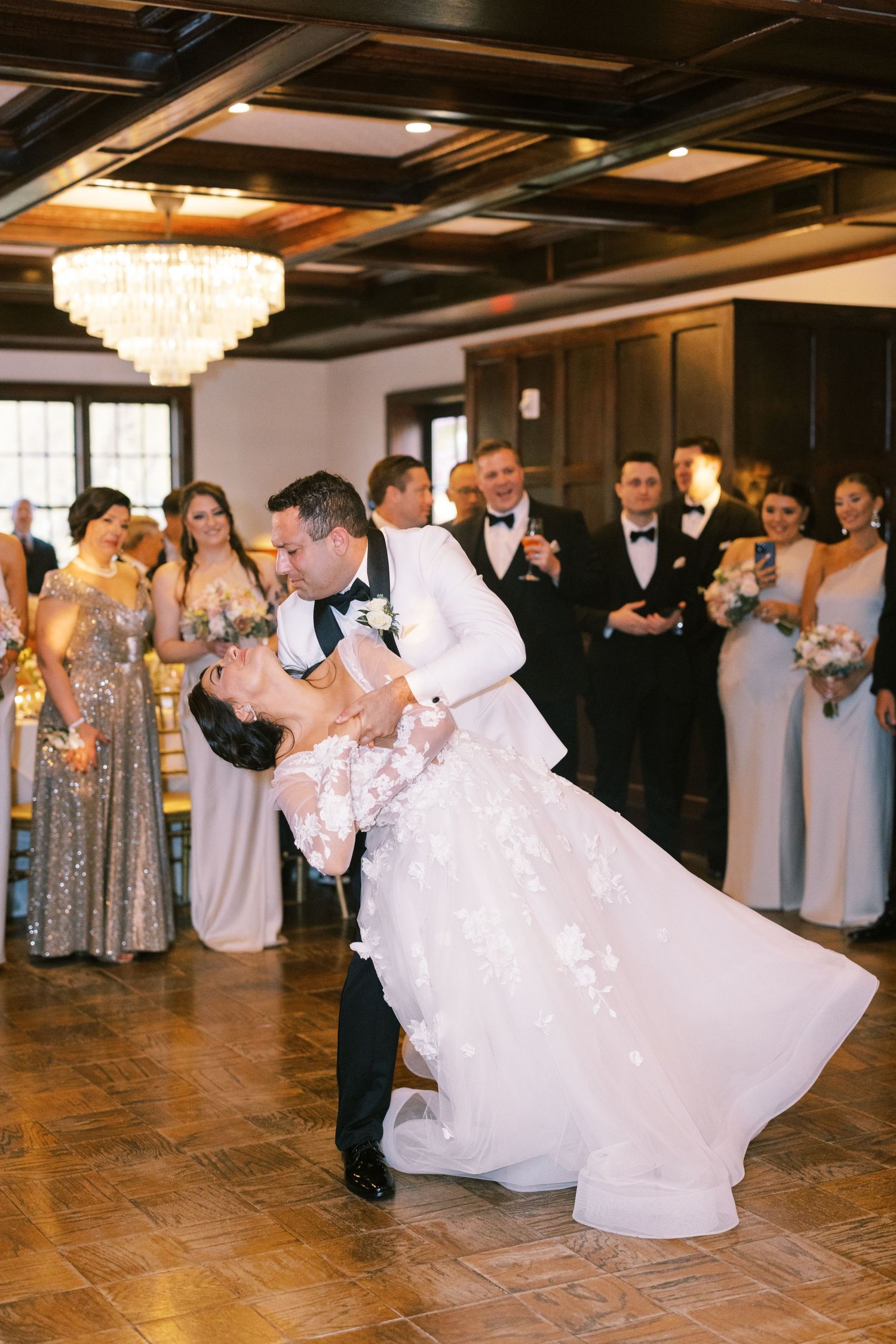 Groom dips bride during first dance surrounded by wedding party in elegant ballroom with chandelier.