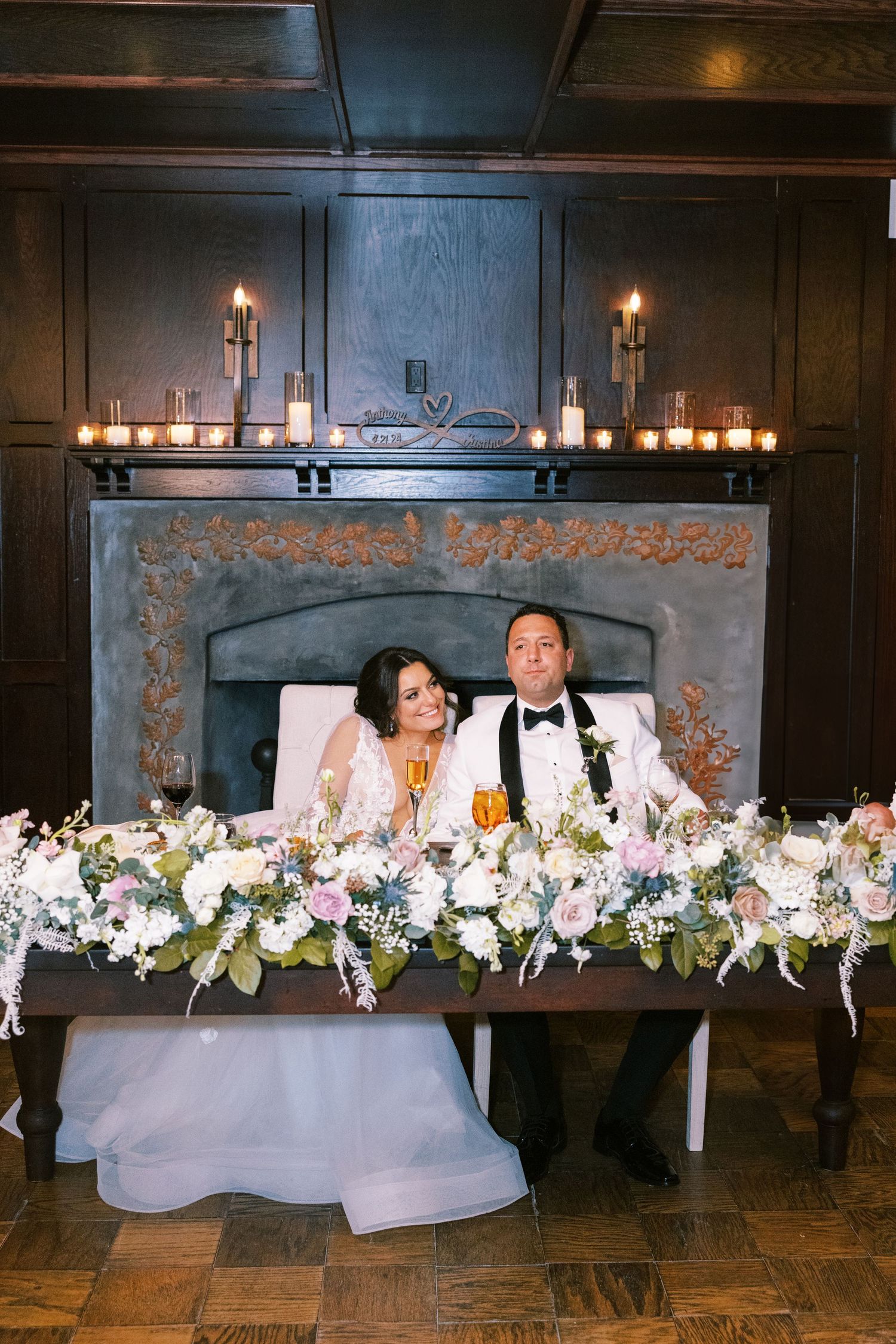 Bride and groom seated at sweetheart table adorned with lush floral garland before lit fireplace.