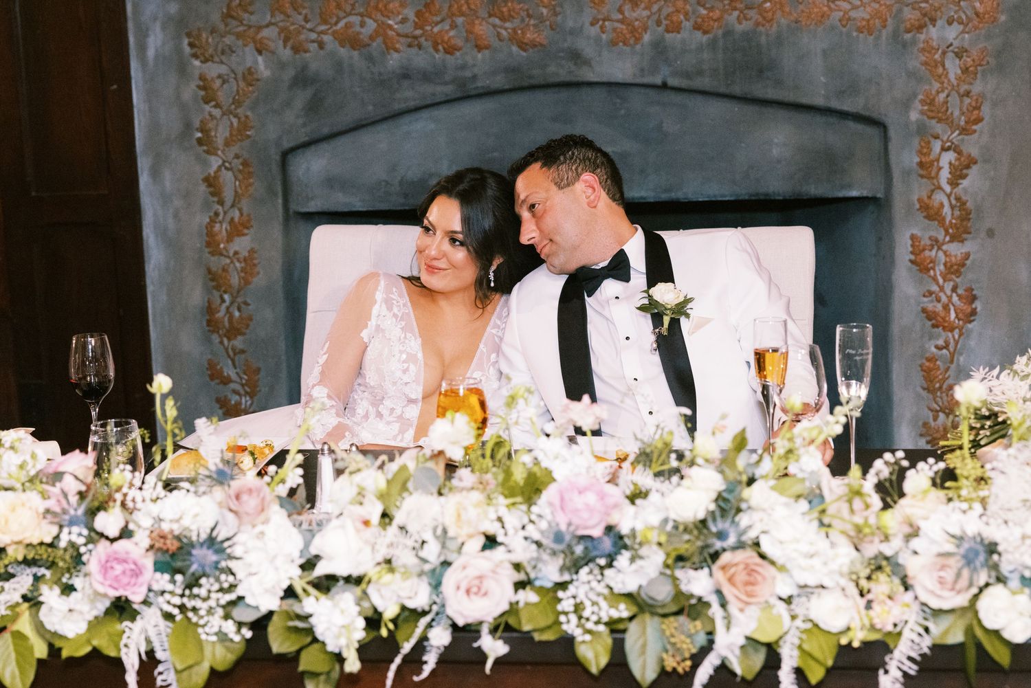 Bride and groom share intimate moment at sweetheart table with pastel floral arrangements and fireplace.
