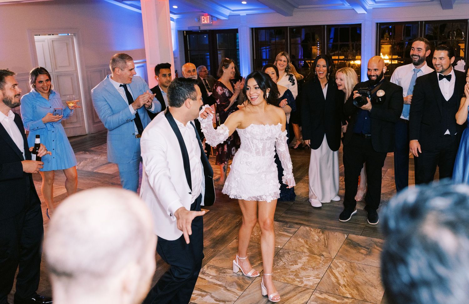 Bride in white mini dress dancing with groom surrounded by cheering guests at wedding reception.