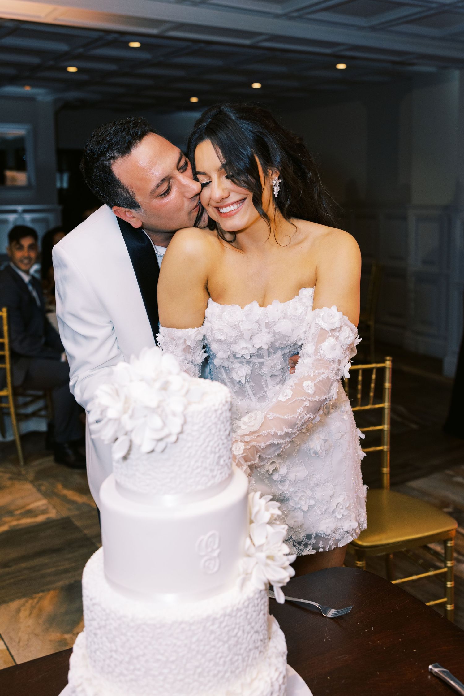 Bride and groom share a kiss while cutting elegant three-tier white wedding cake at reception.