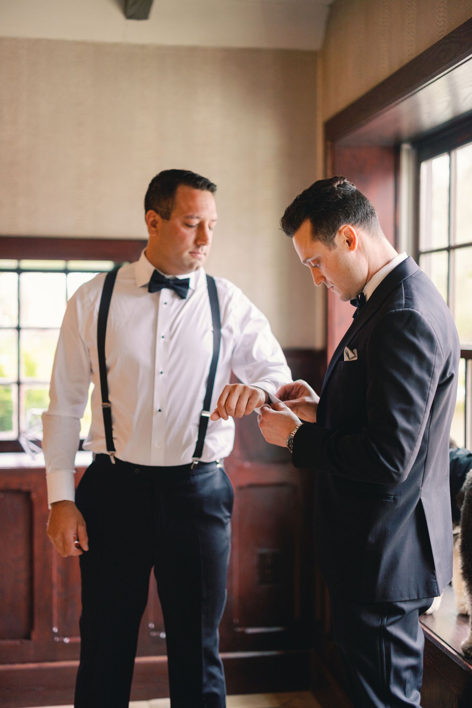 Groom getting cufflinks fastened by groomsman in white shirt with navy suspenders and bow tie before wedding.