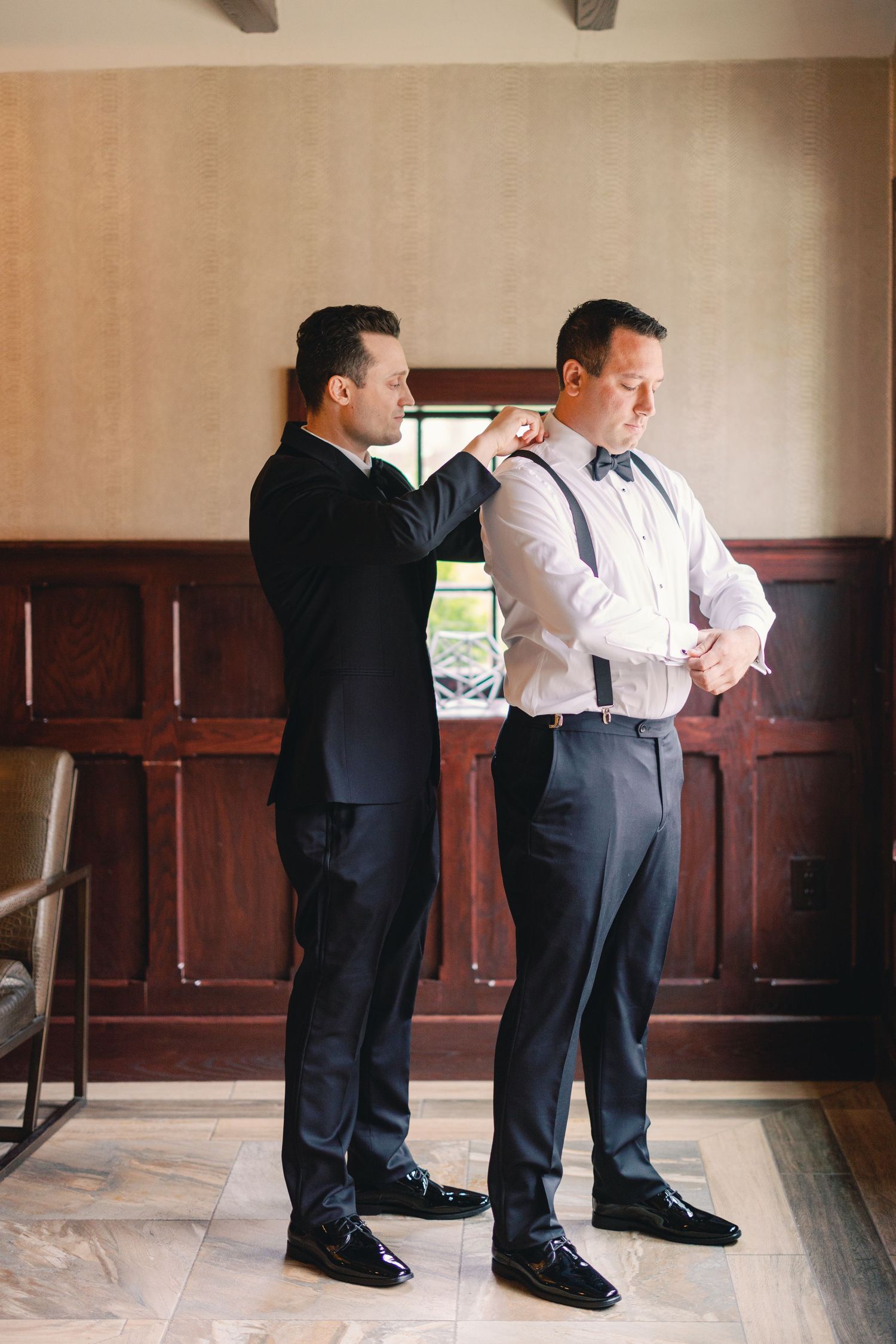 Groomsman helping groom adjust suspenders while getting ready for wedding in wood-paneled dressing room.