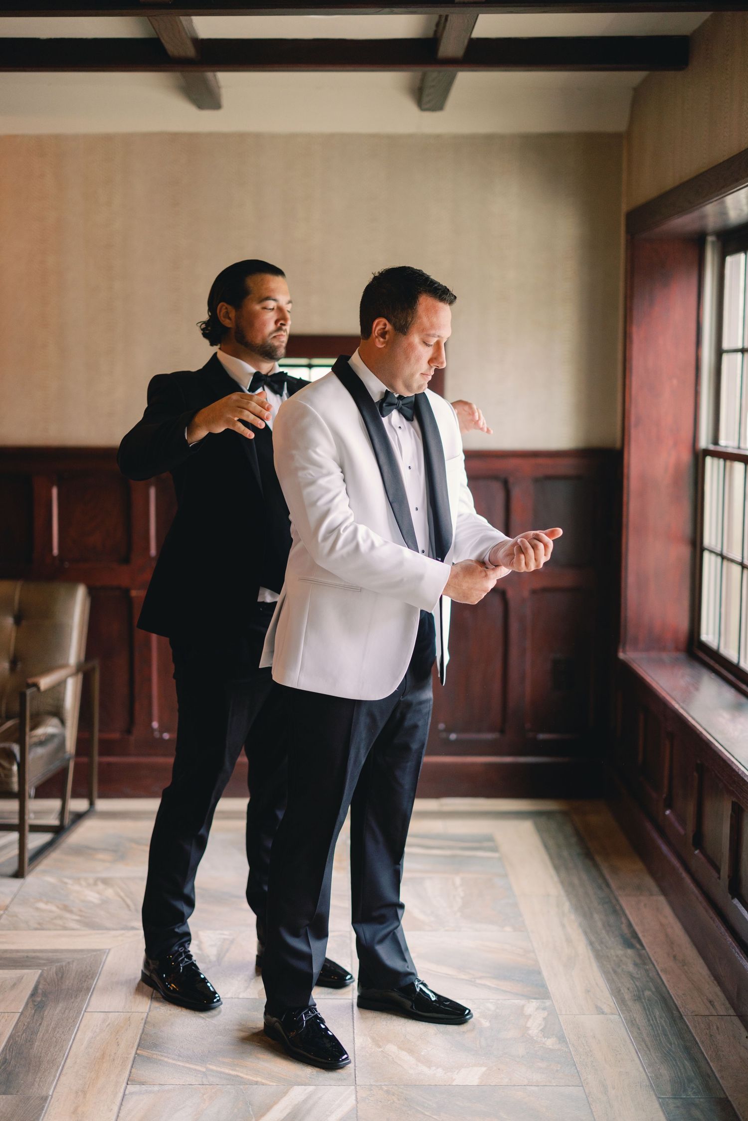 Two groomsmen getting ready indoors, one in white jacket checking cufflinks, other adjusting bow tie.