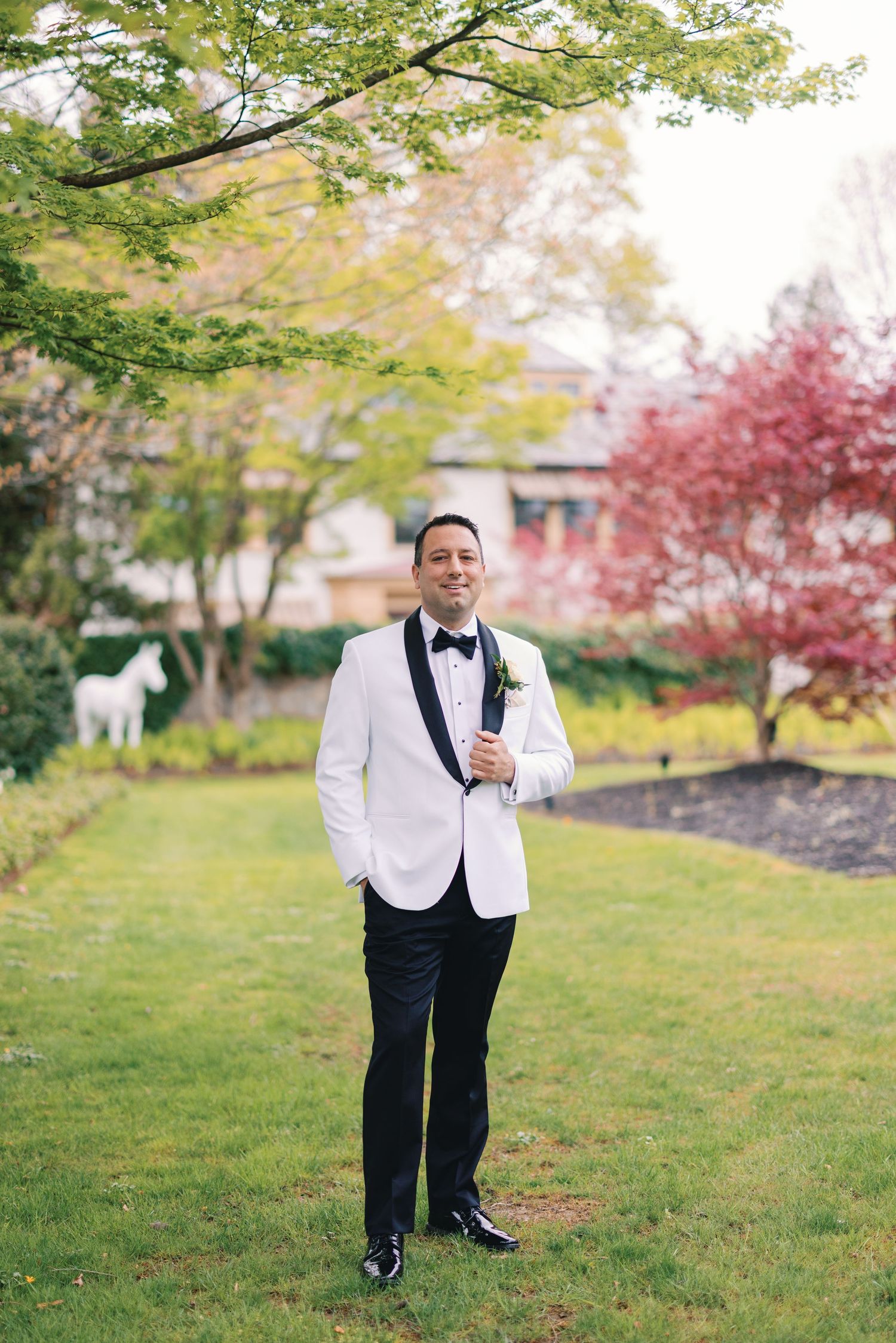 Dapper groom in white tuxedo jacket with black lapels and boutonniere posing in lush garden setting.