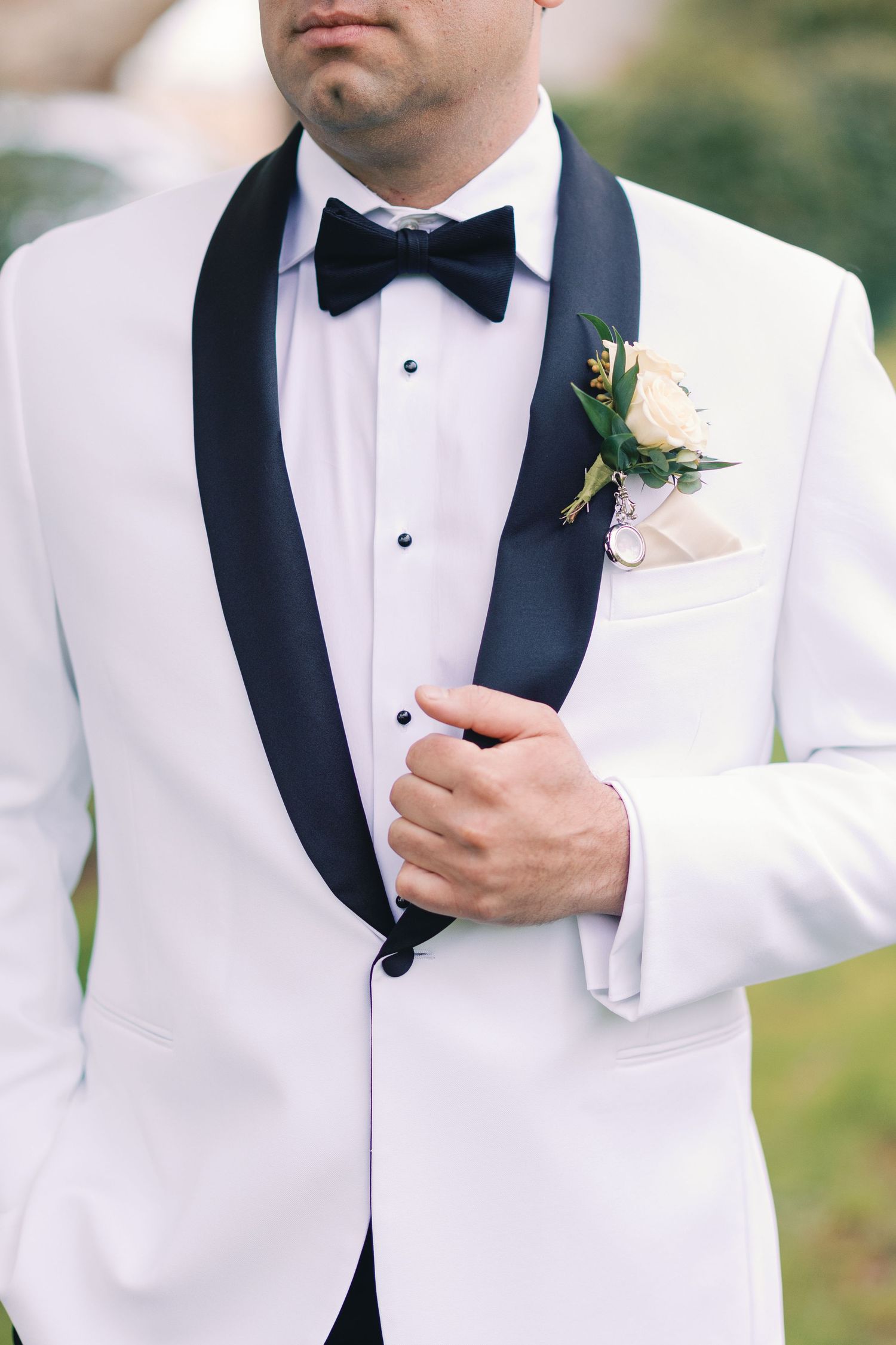 Close-up of groom in white tuxedo with black shawl lapels, bow tie, and floral boutonniere on lapel.