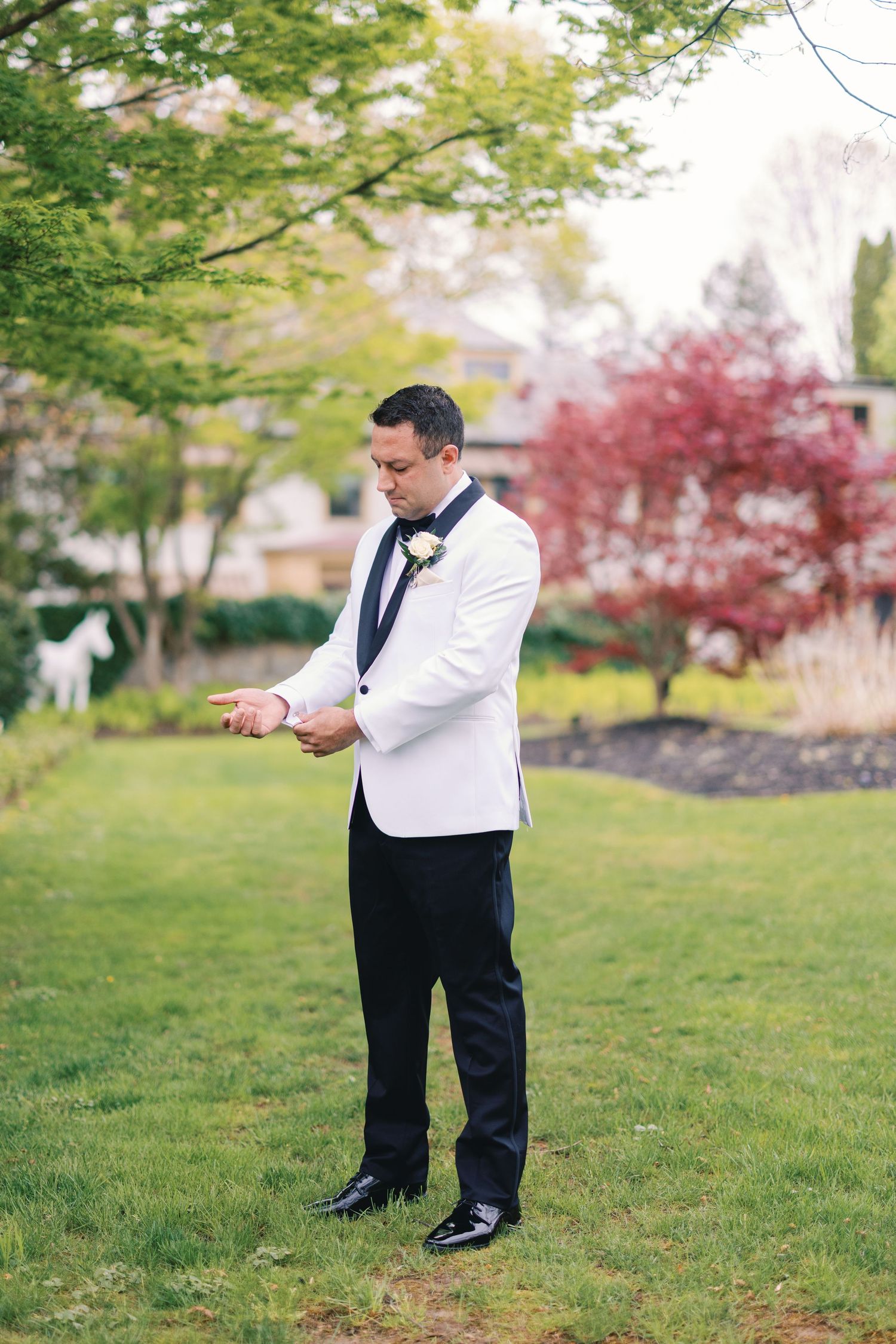 Groom in white tuxedo jacket with black lapels checking cufflinks while standing on green garden lawn.