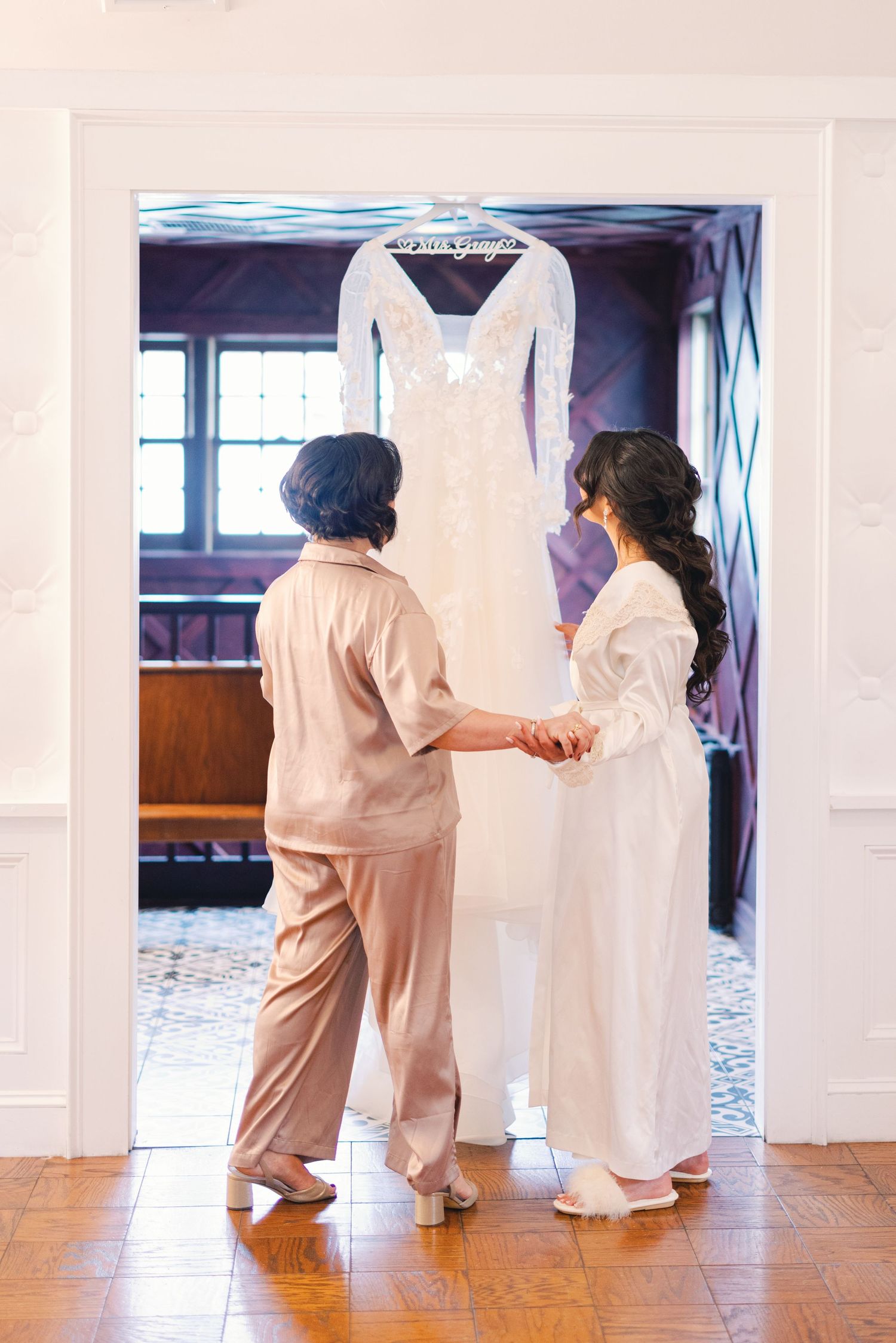 Bride and mother holding hands while admiring wedding gown hanging in doorway before ceremony.