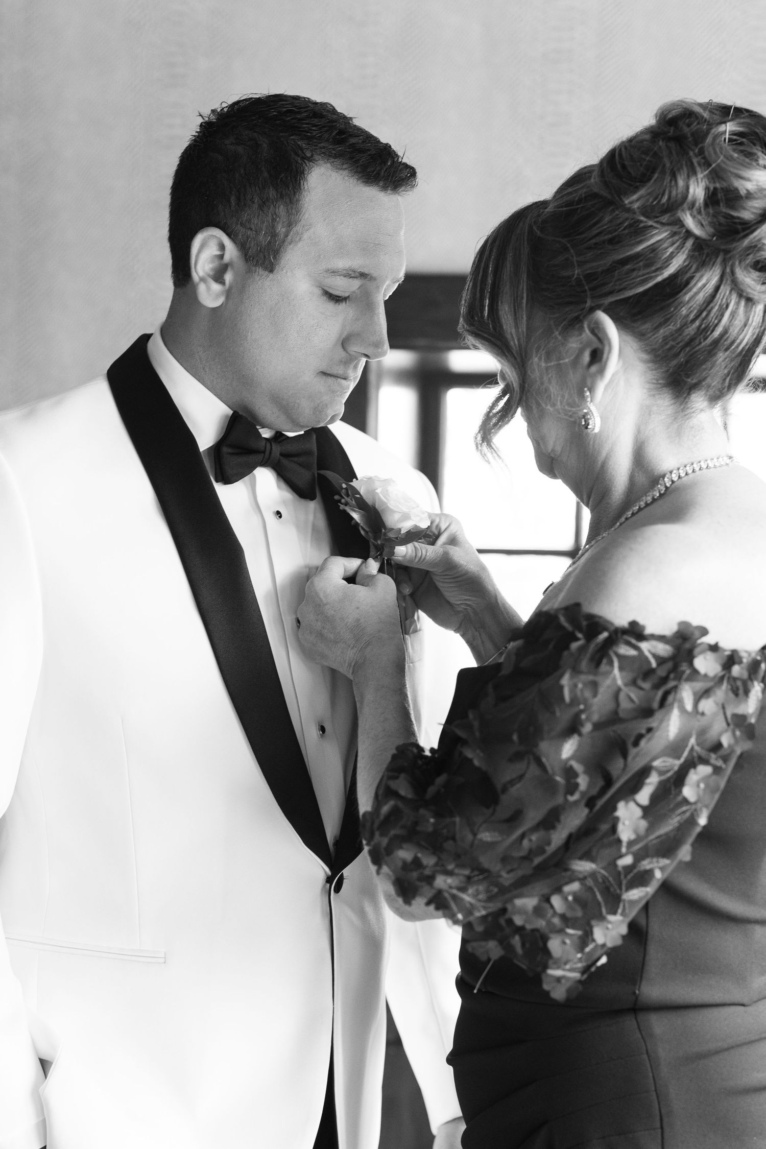 Black and white photo of woman pinning boutonniere on groom in white tuxedo before wedding ceremony.