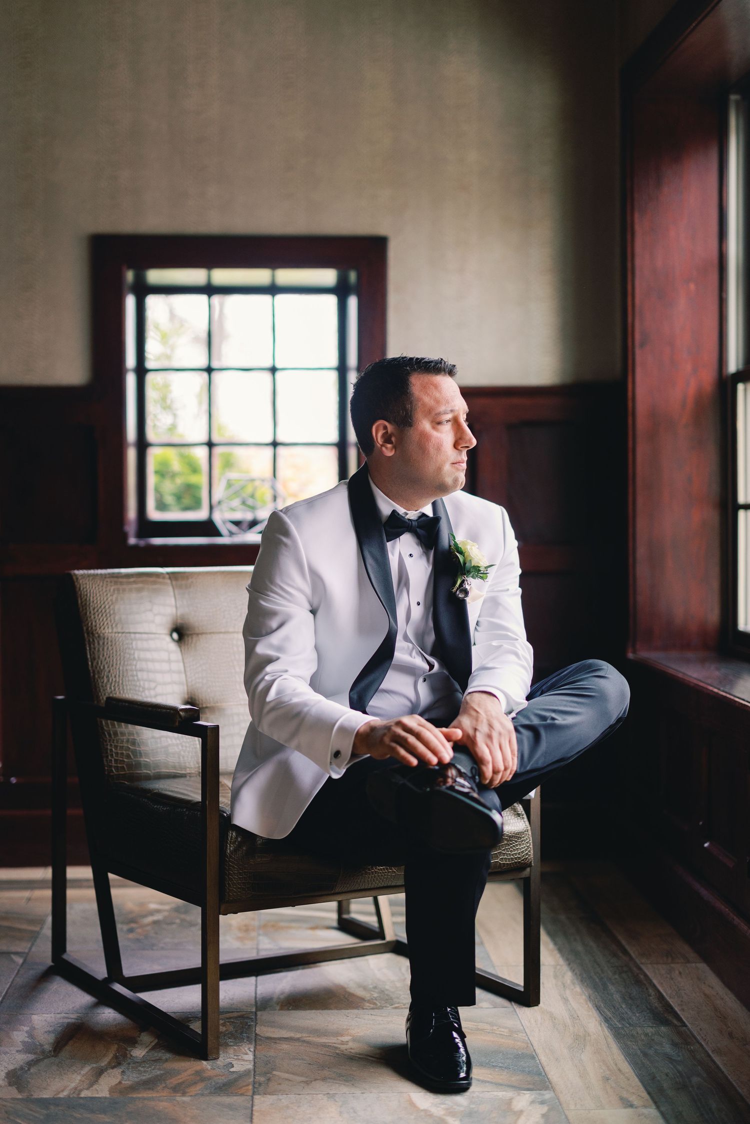 Groom in white tuxedo with black bow tie sits contemplatively in a chair by a window on his wedding day.