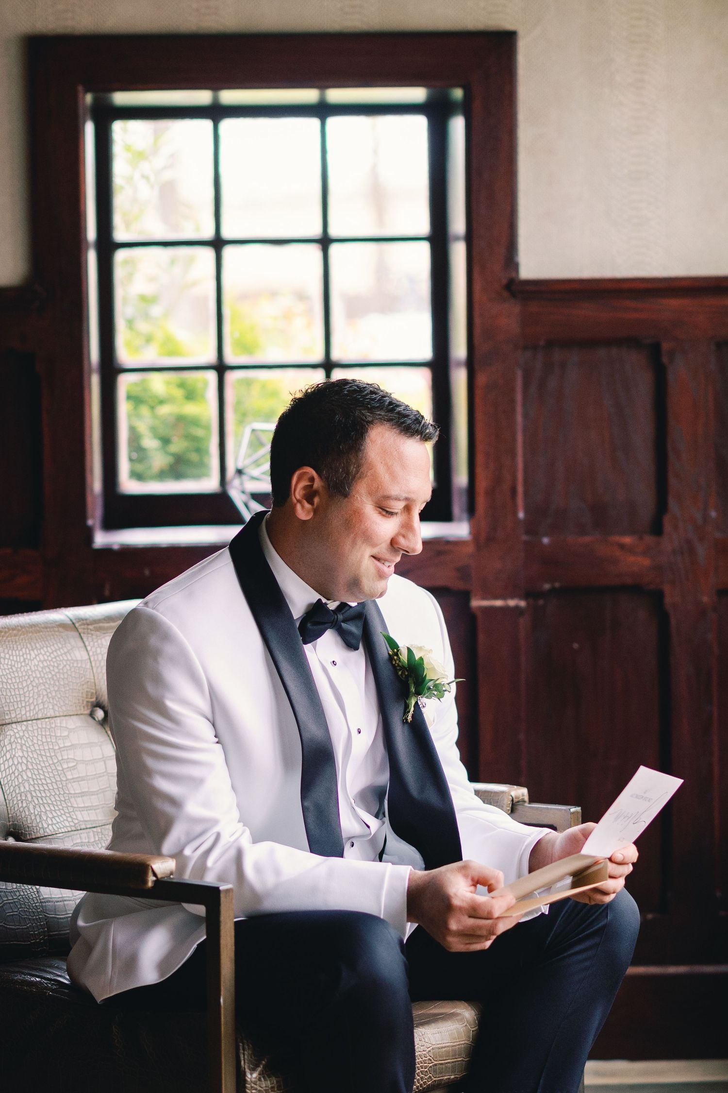 Groom in white tuxedo jacket reads a heartfelt letter while seated in an elegant chair before his wedding.
