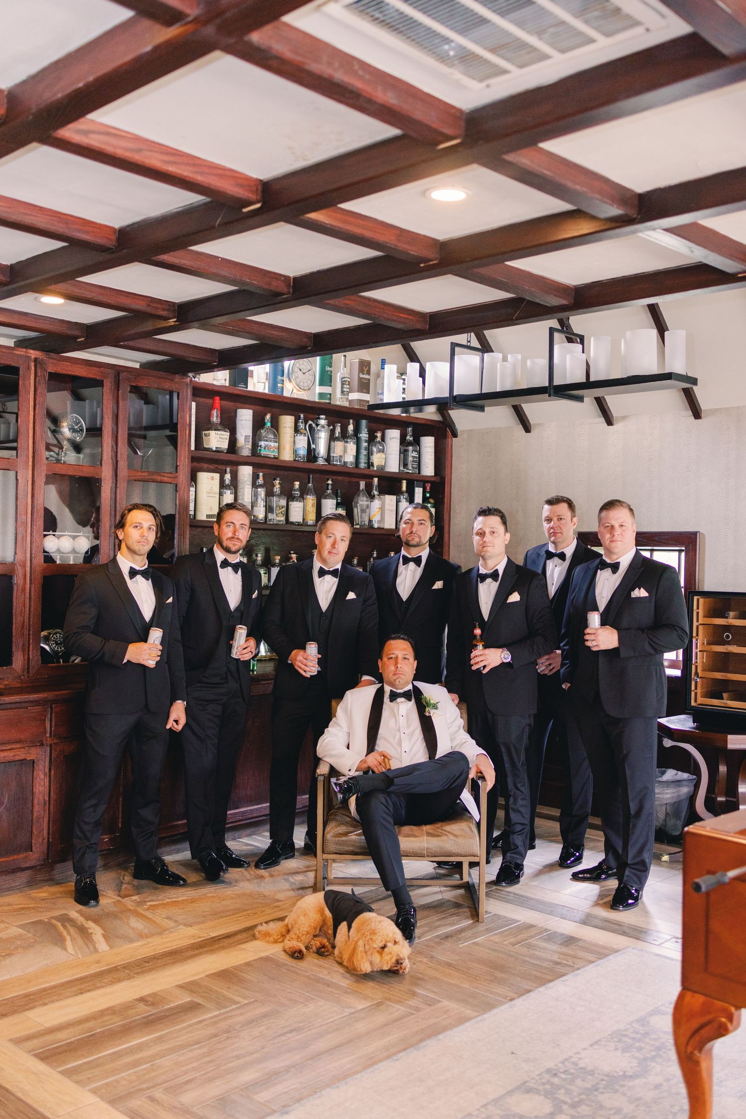 Groom and groomsmen in black tuxedos pose together in a bar with a golden retriever on the floor.