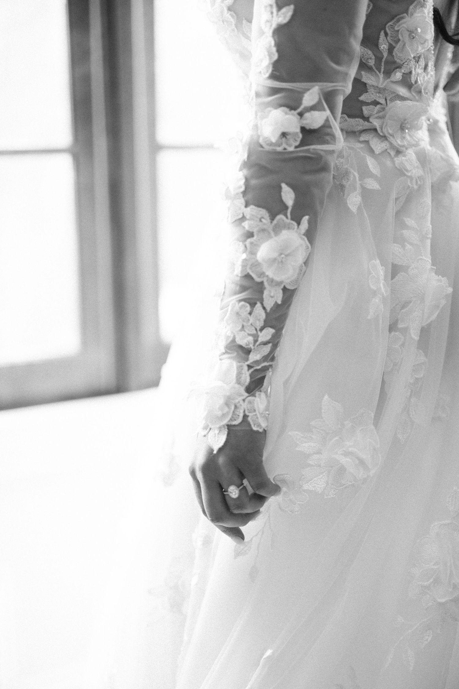Close-up black and white detail of a bride's floral lace wedding gown sleeve with engagement ring visible.
