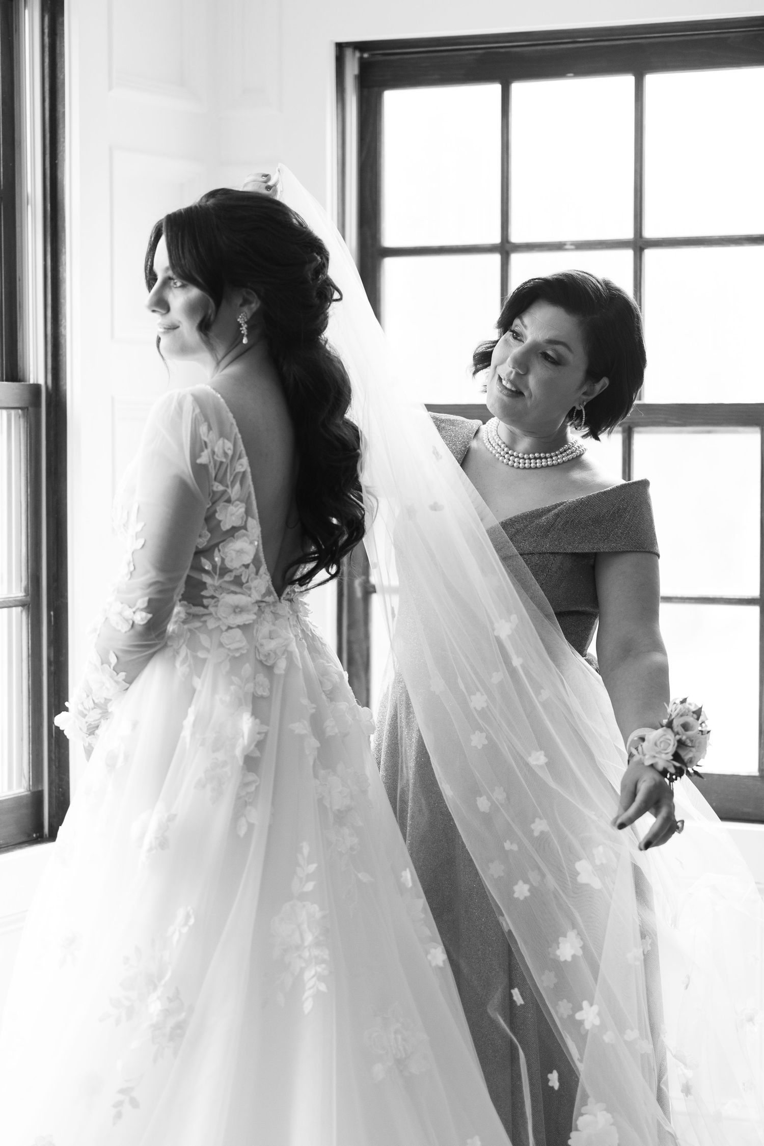 Black and white photo of a bride in floral lace gown with veil being adjusted by her mother near a window.