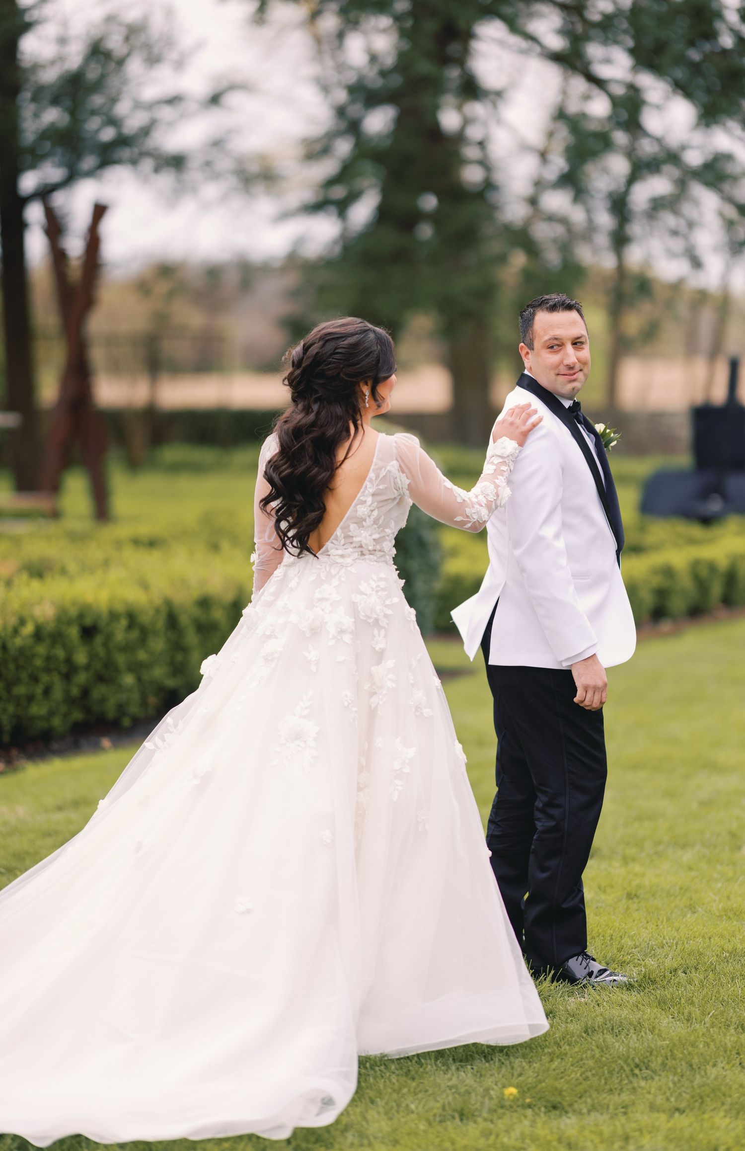 Bride in floral lace backless gown taps groom's shoulder during outdoor first look in a lush garden.