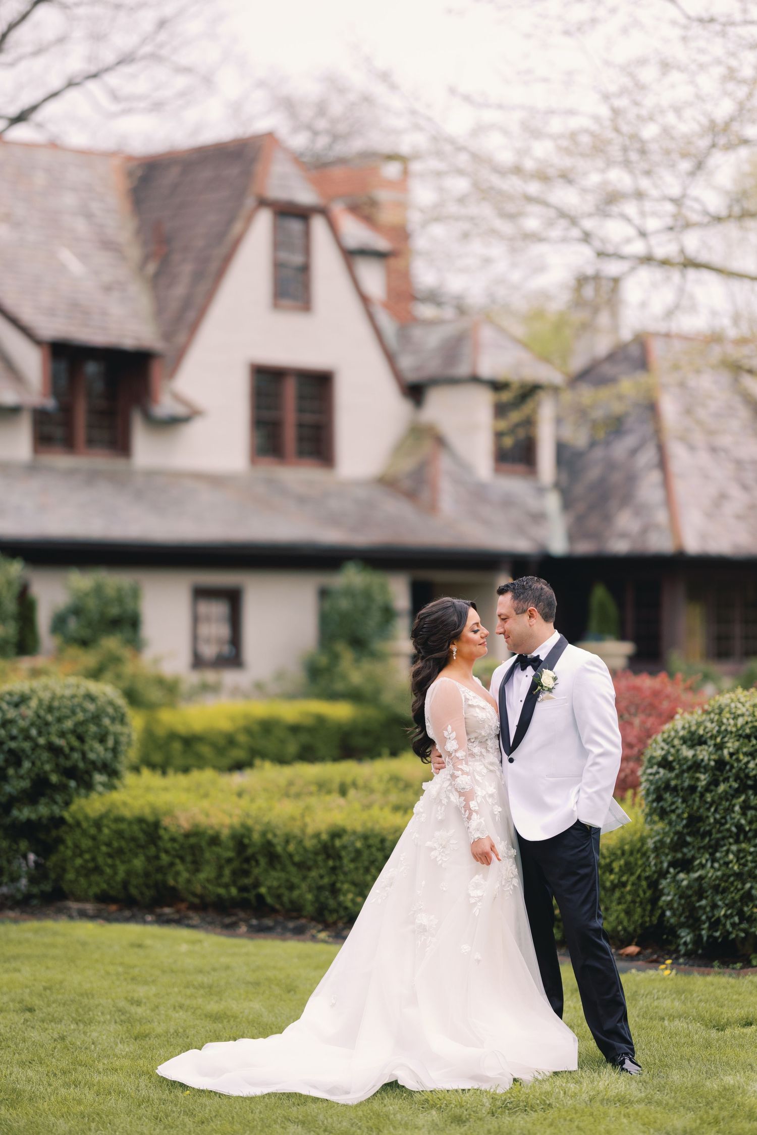 Bride and groom share a romantic moment in a manicured garden with a Tudor-style estate in the background.