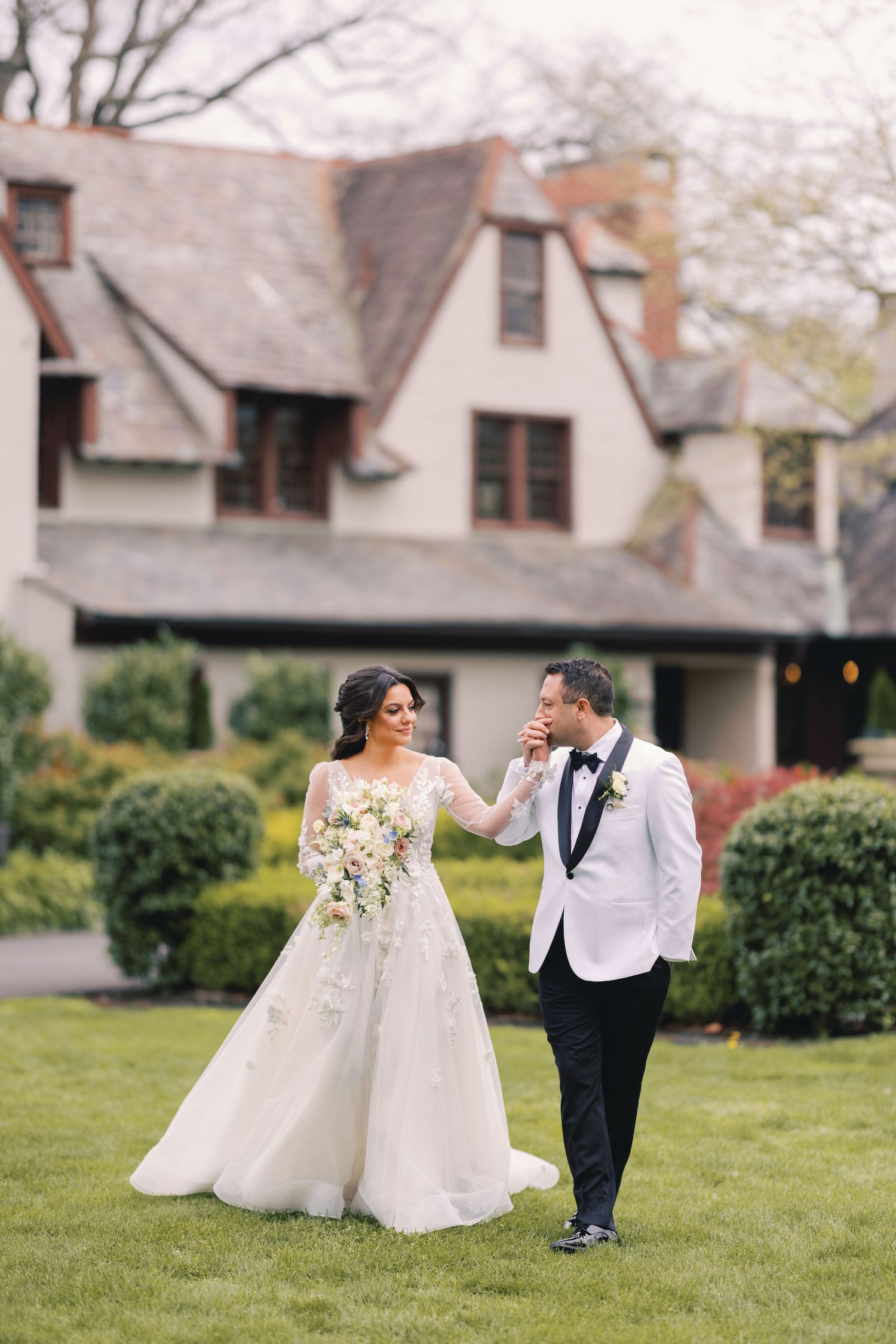 Bride and groom walk through a garden as the groom kisses her hand, with a Tudor mansion behind them.