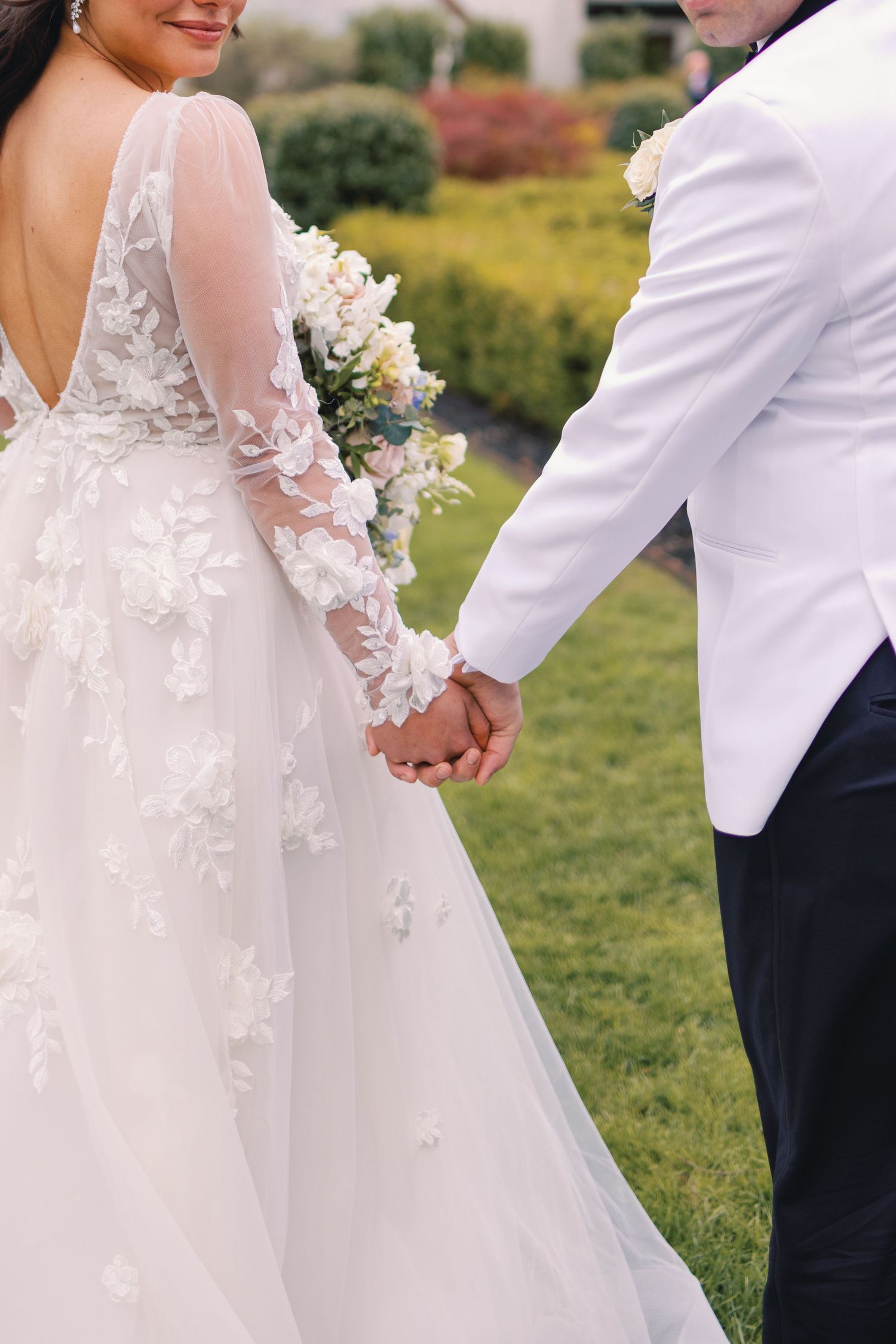 Bride and groom hold hands outdoors, showcasing the bride's floral lace gown and white bouquet.