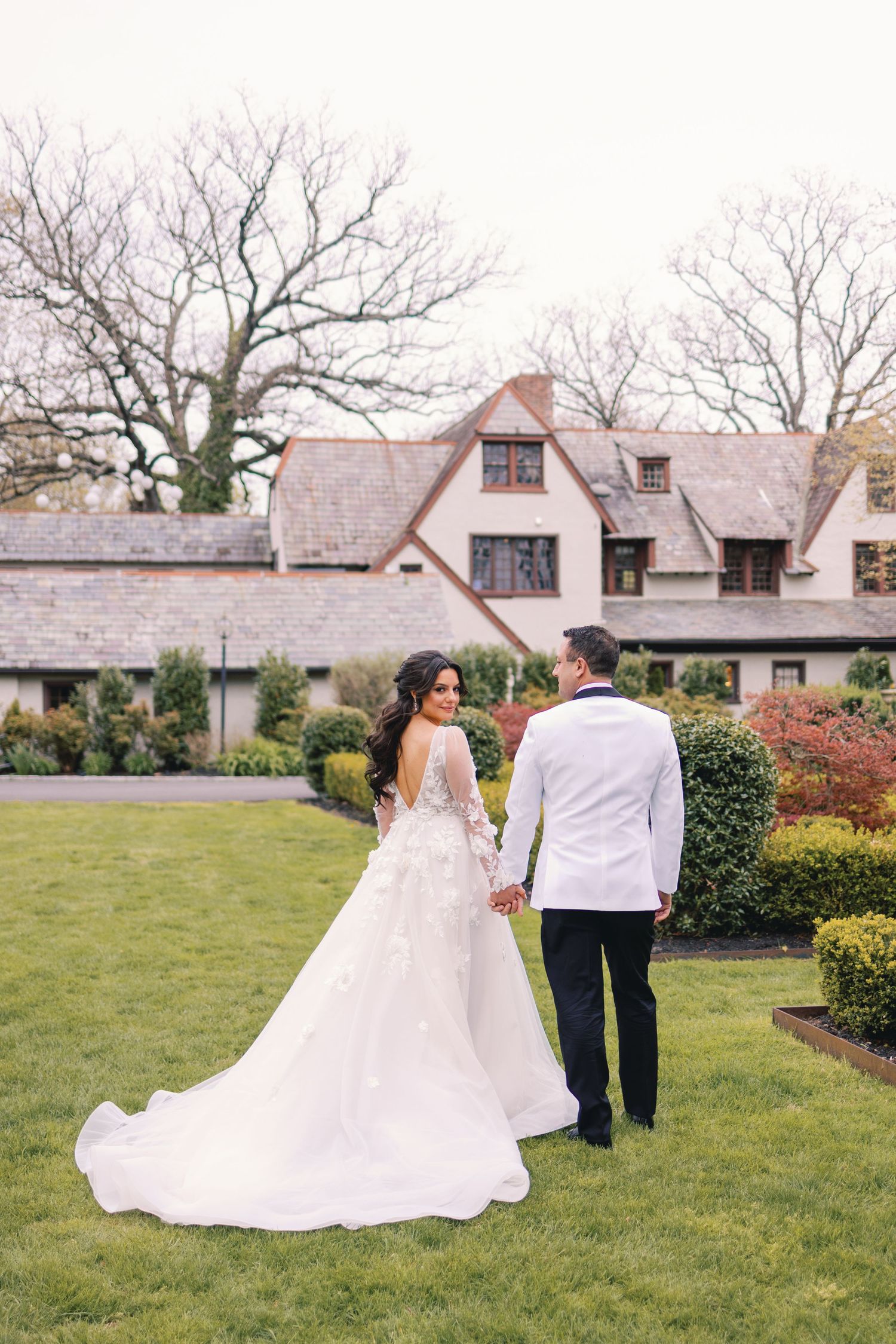 Bride in floral ballgown and groom in white tuxedo hold hands walking toward elegant estate venue.