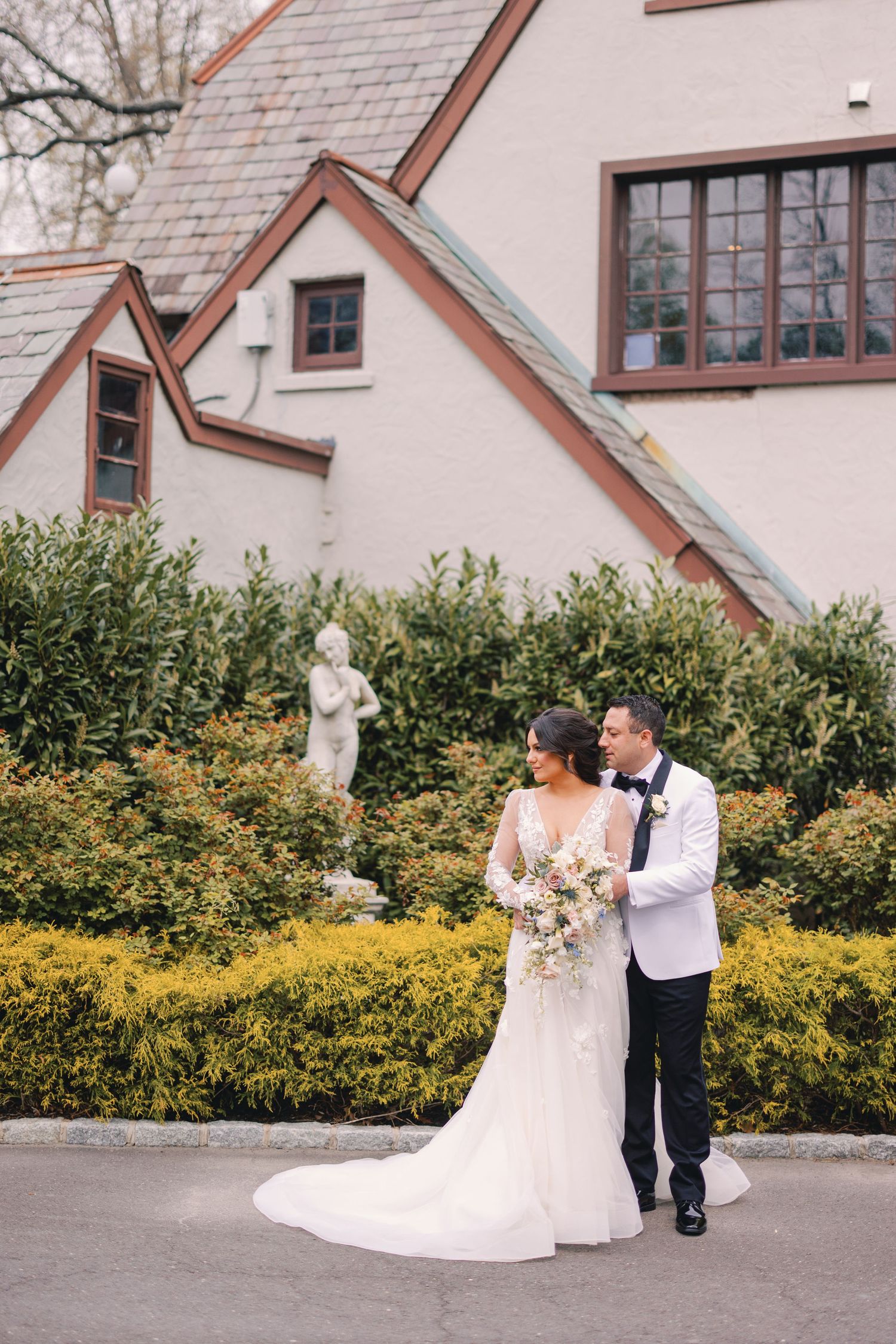 Bride and groom walk together near a white estate surrounded by lush hedges and garden statuary.