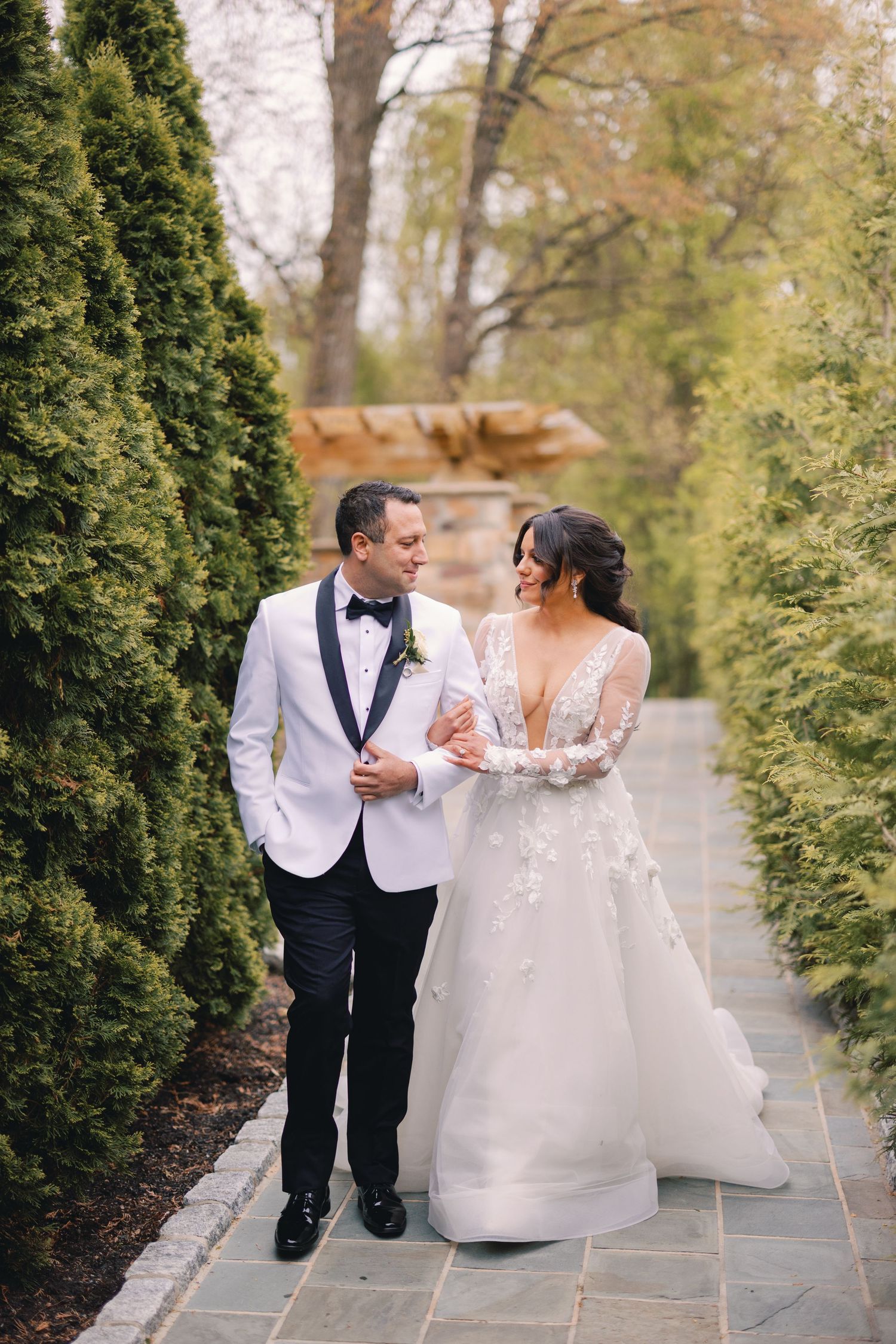 Bride and groom in white tuxedo walk arm in arm along lush green hedge-lined garden pathway.