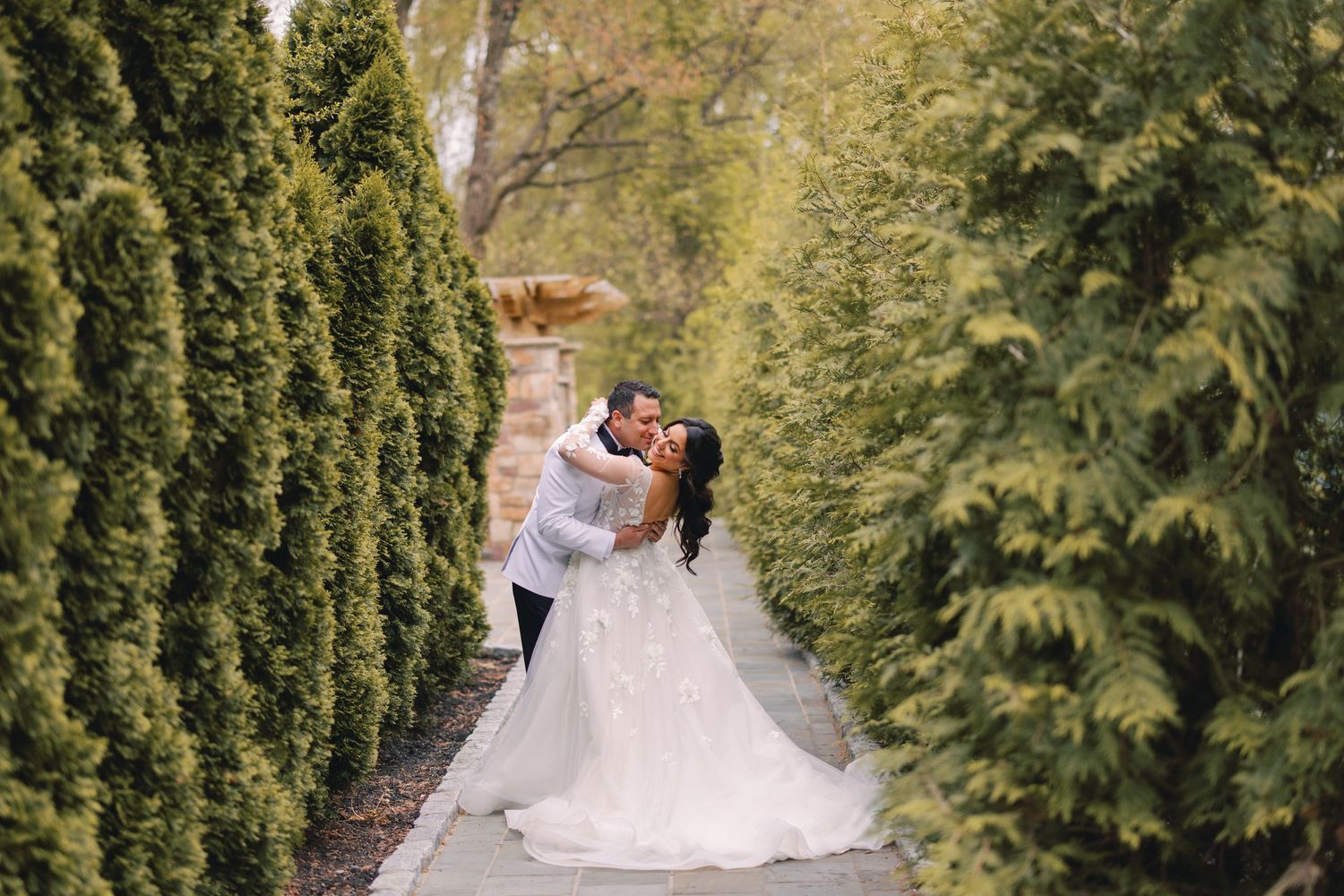 Bride and groom share a passionate kiss along a tree-lined garden pathway at their wedding.