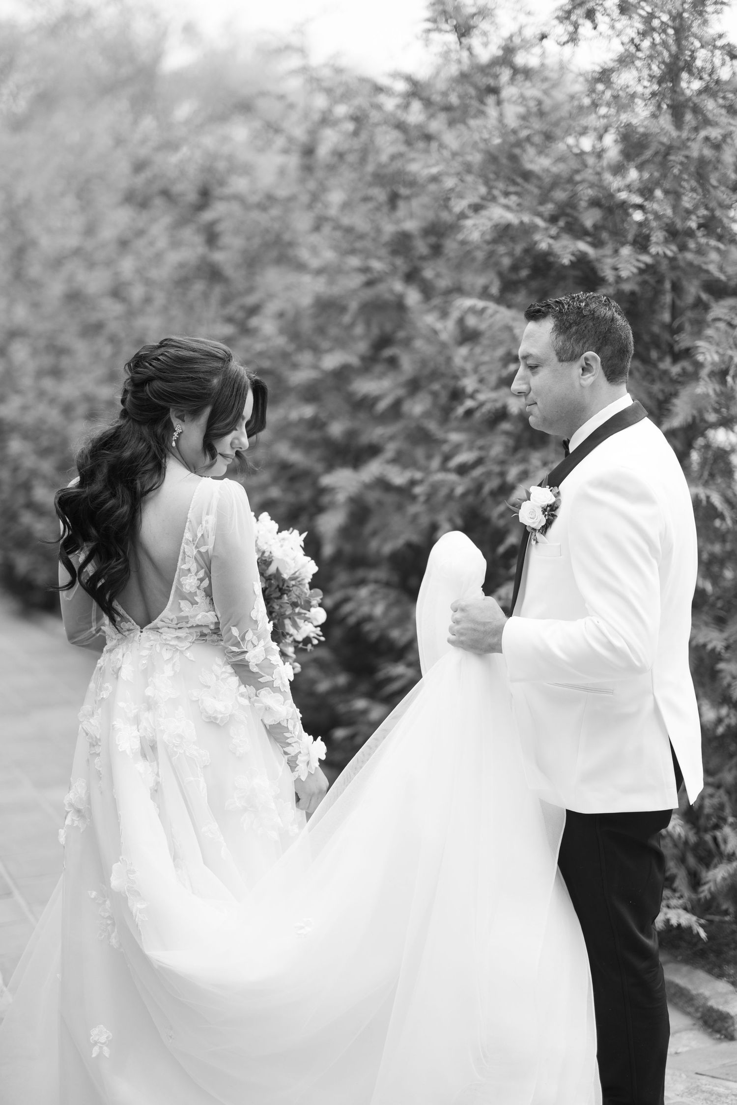 Black and white photo of bride in backless floral gown and groom sharing romantic moment outdoors.