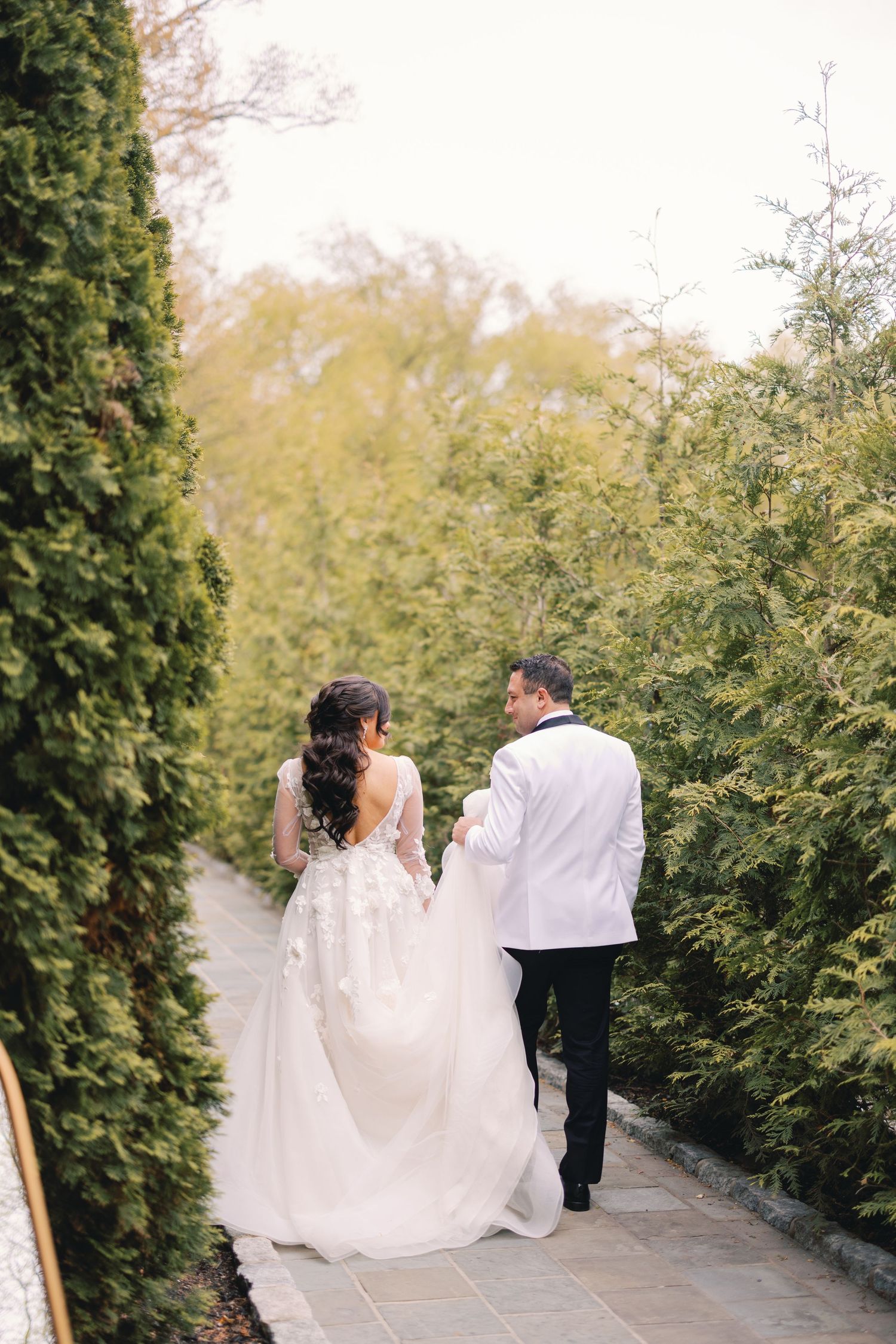 Bride in lace ballgown and groom in white tuxedo walk hand in hand through green hedged path.