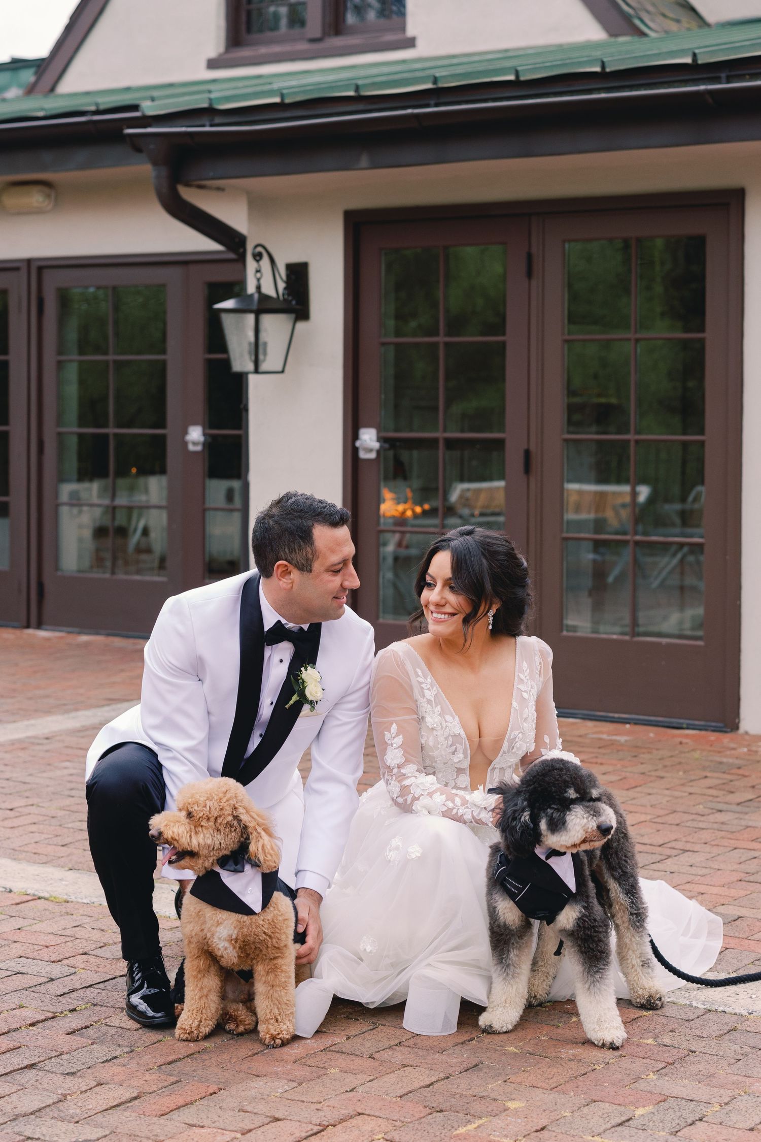 Smiling bride and groom in white tuxedo pose with two dogs wearing bow ties on brick patio.