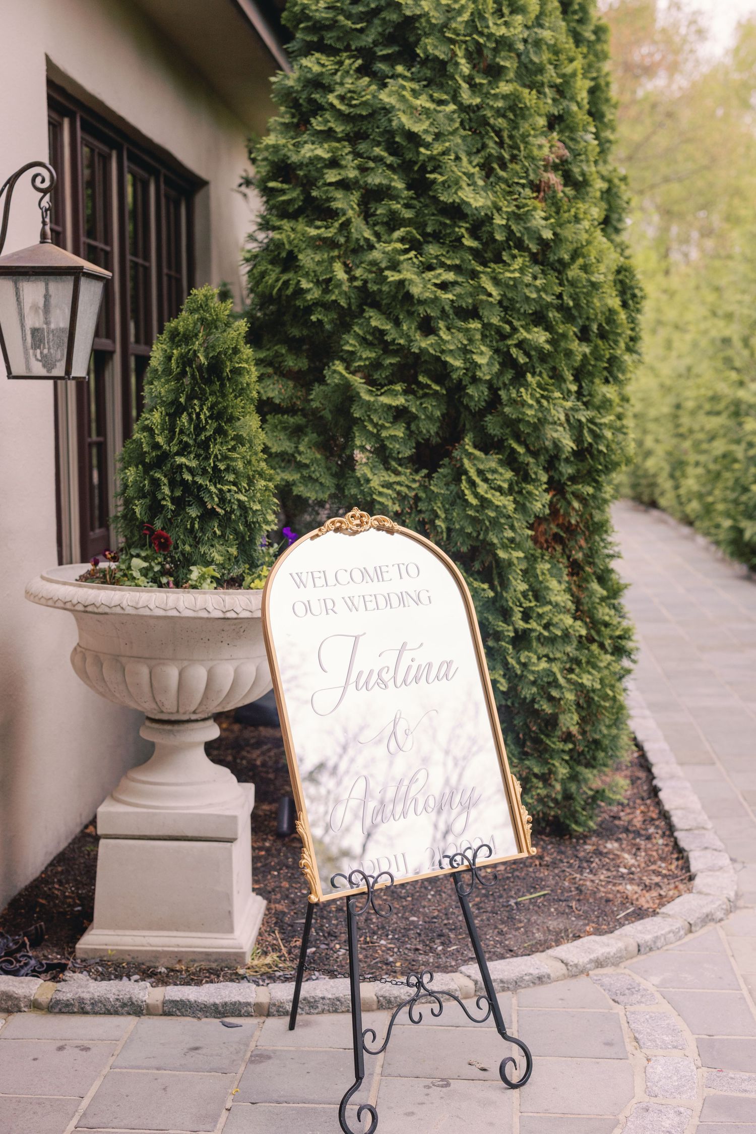 Gold-framed wedding welcome mirror sign for Justina and Johnny displayed on black easel outside elegant venue.