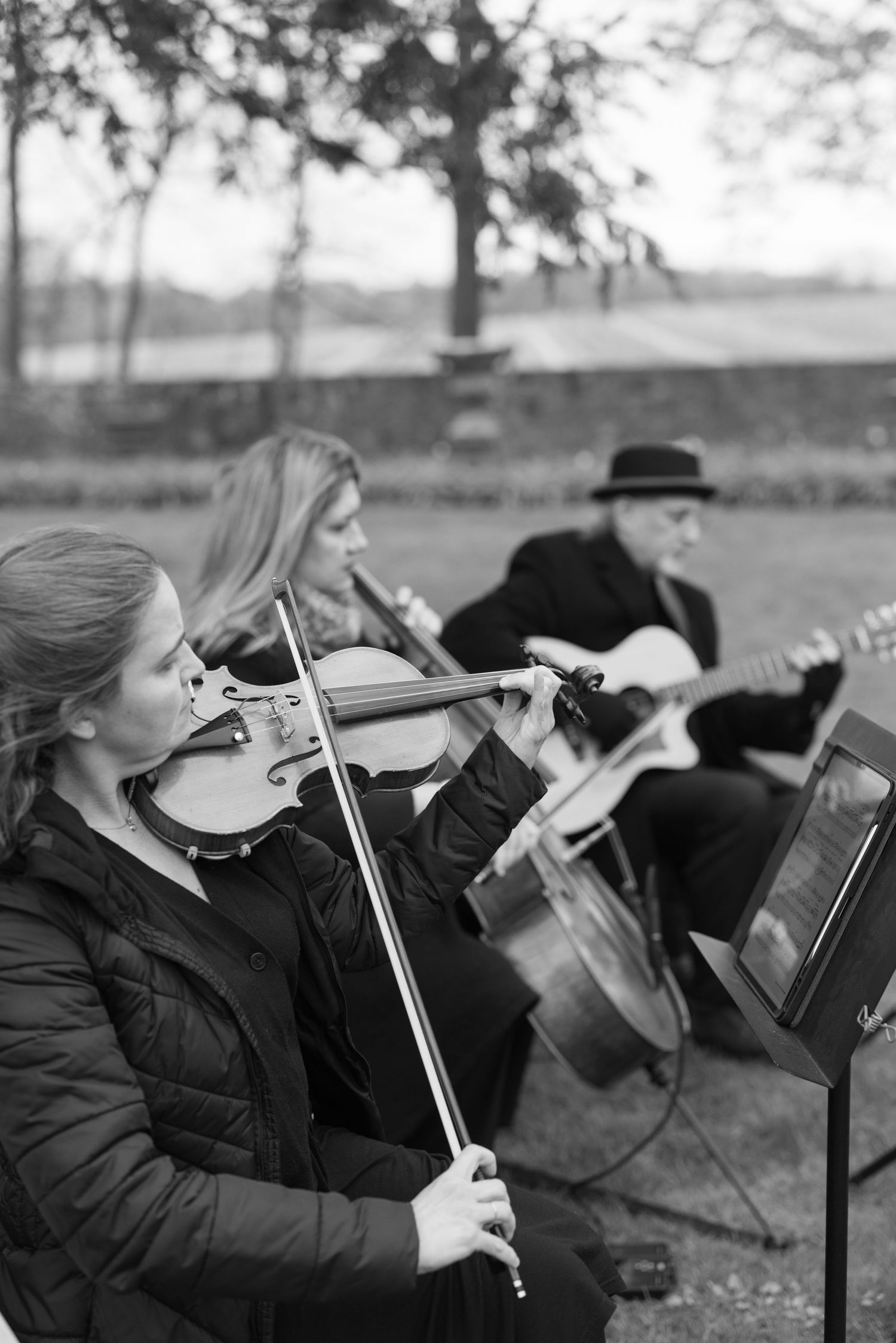 Black and white photo of string musicians playing violin and cello at outdoor wedding ceremony.