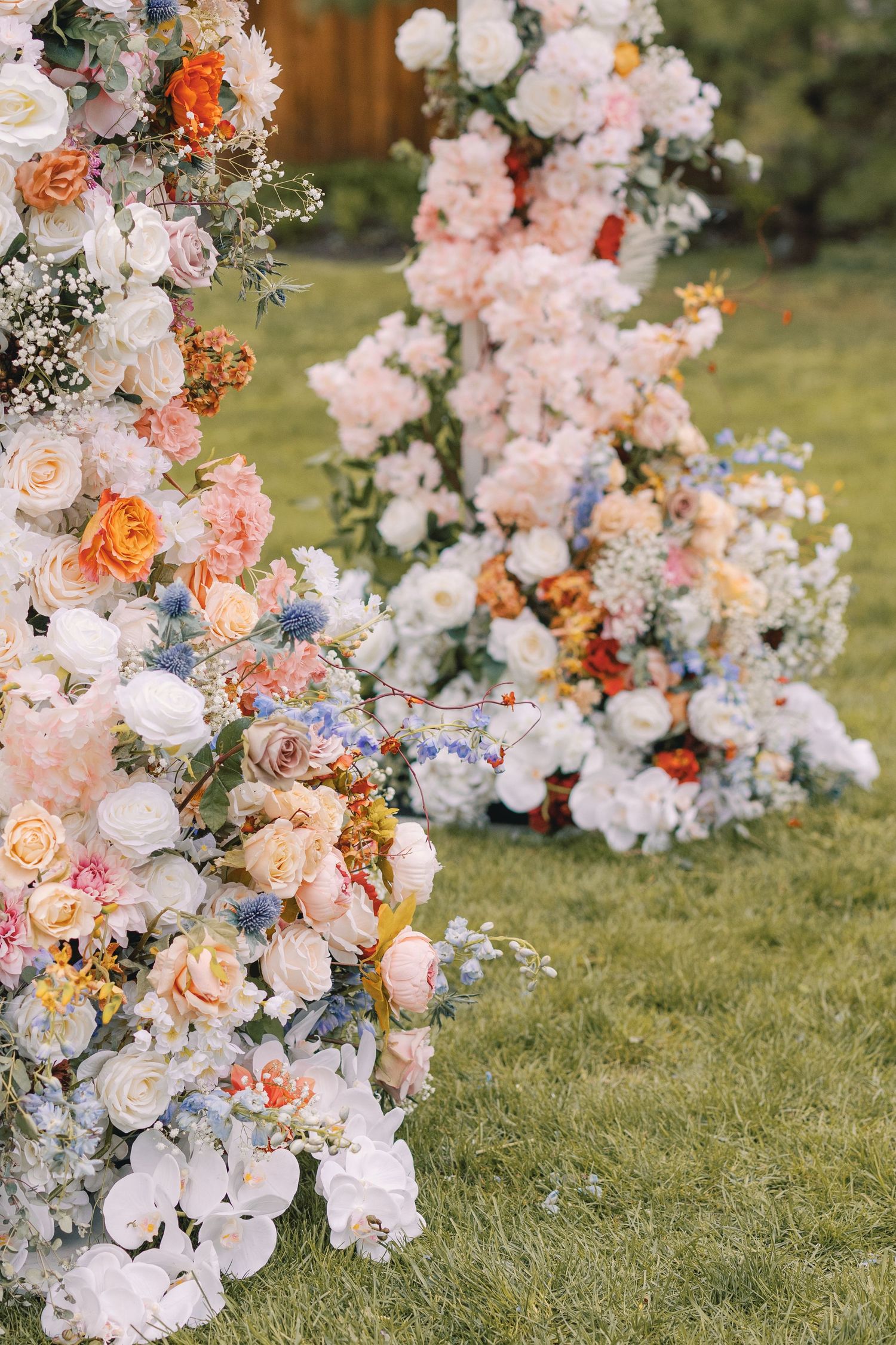 Lush colorful wedding ceremony floral arch with pink, white, orange and blue flowers on green lawn.