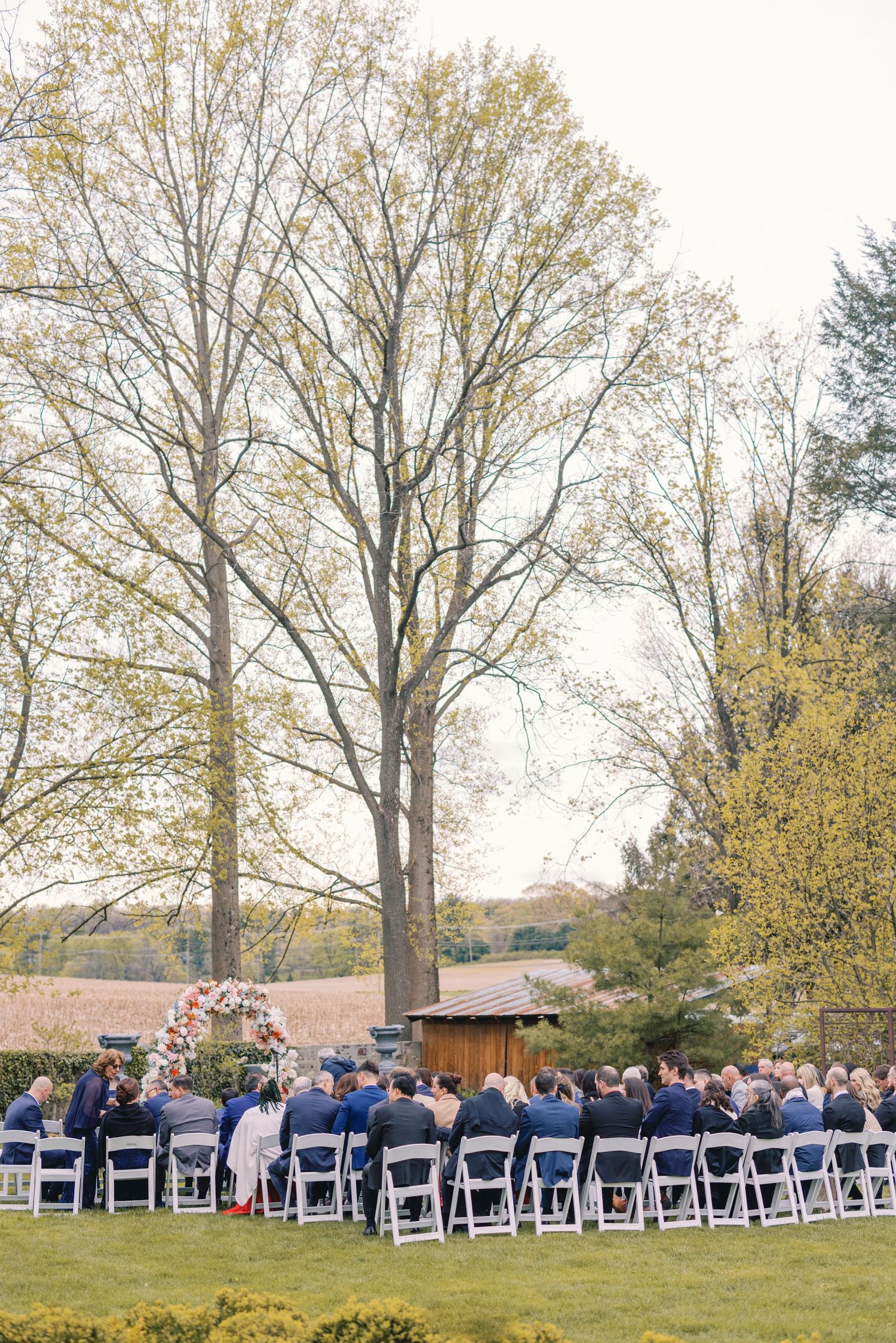 Outdoor wedding ceremony with guests seated in white chairs beneath tall trees in garden setting.