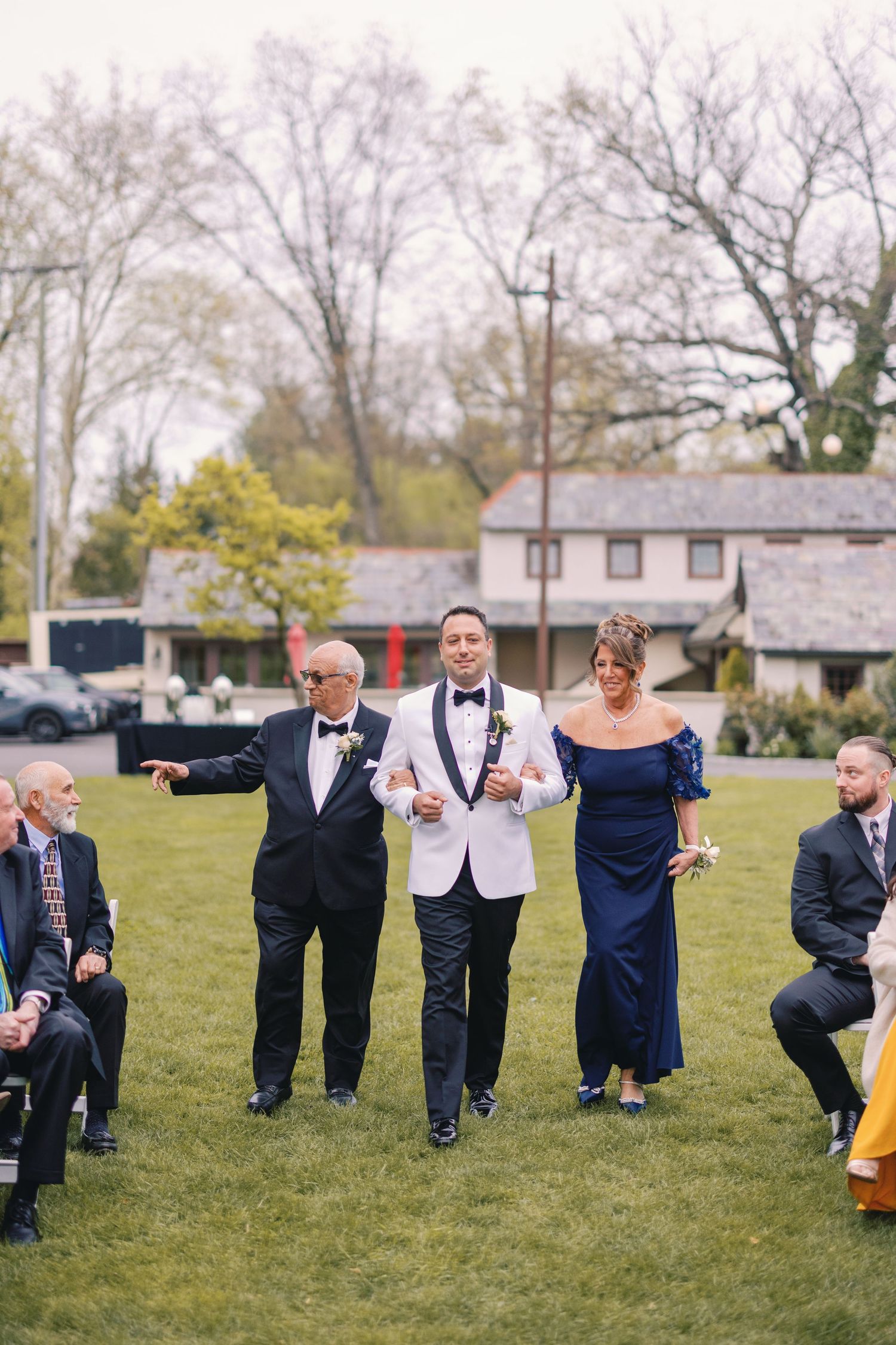 Groom in white tuxedo jacket walks down aisle escorted by parents at outdoor wedding ceremony.