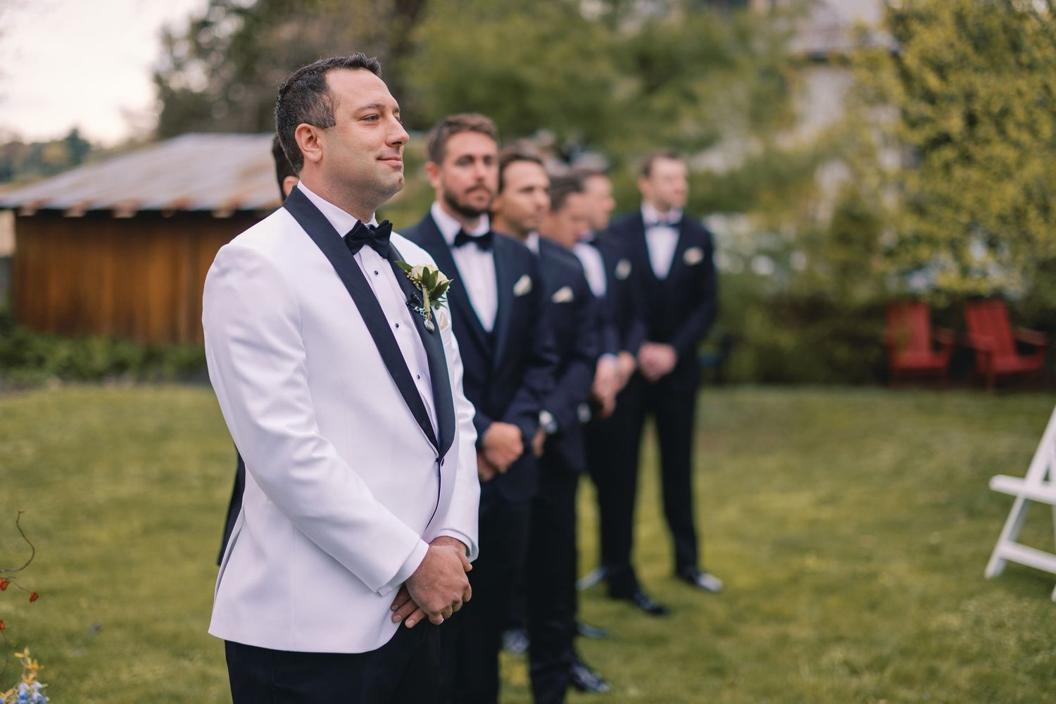Groom in white tuxedo watches bride approach at outdoor wedding ceremony with groomsmen behind him.