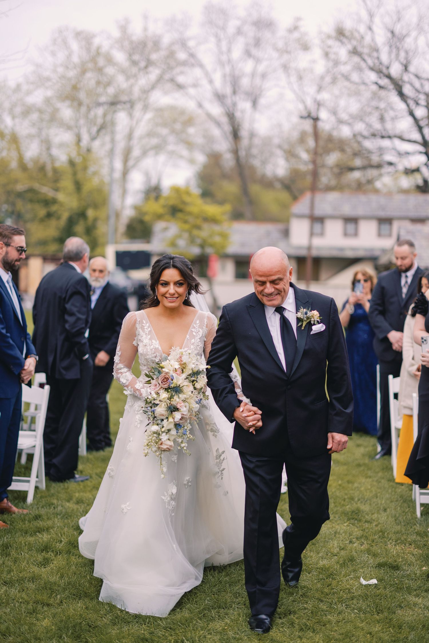 Bride in lace wedding gown holding floral bouquet walks down aisle with father at outdoor ceremony.