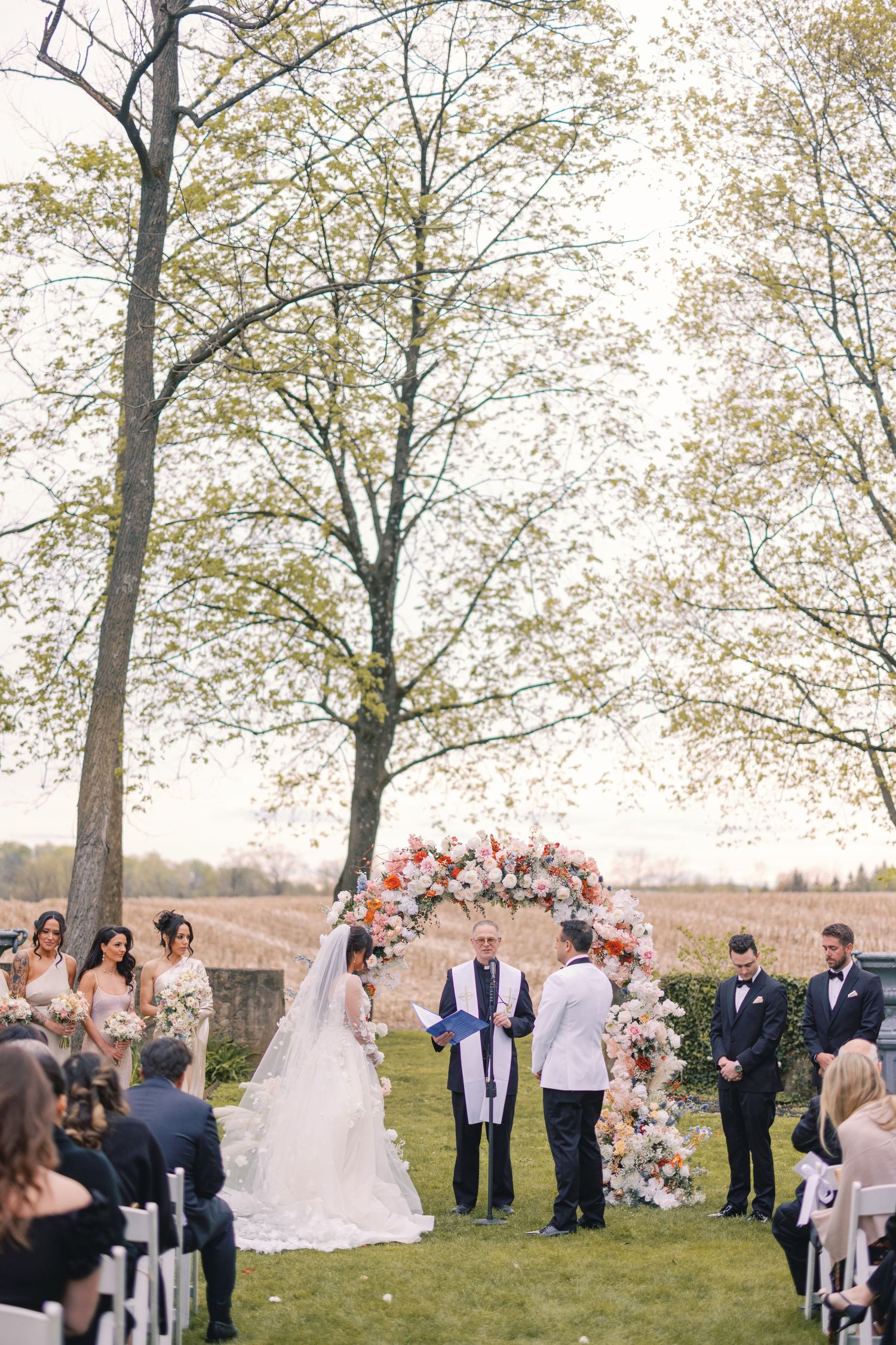 Bride and groom exchange vows at floral arch altar during outdoor wedding ceremony beside open field.